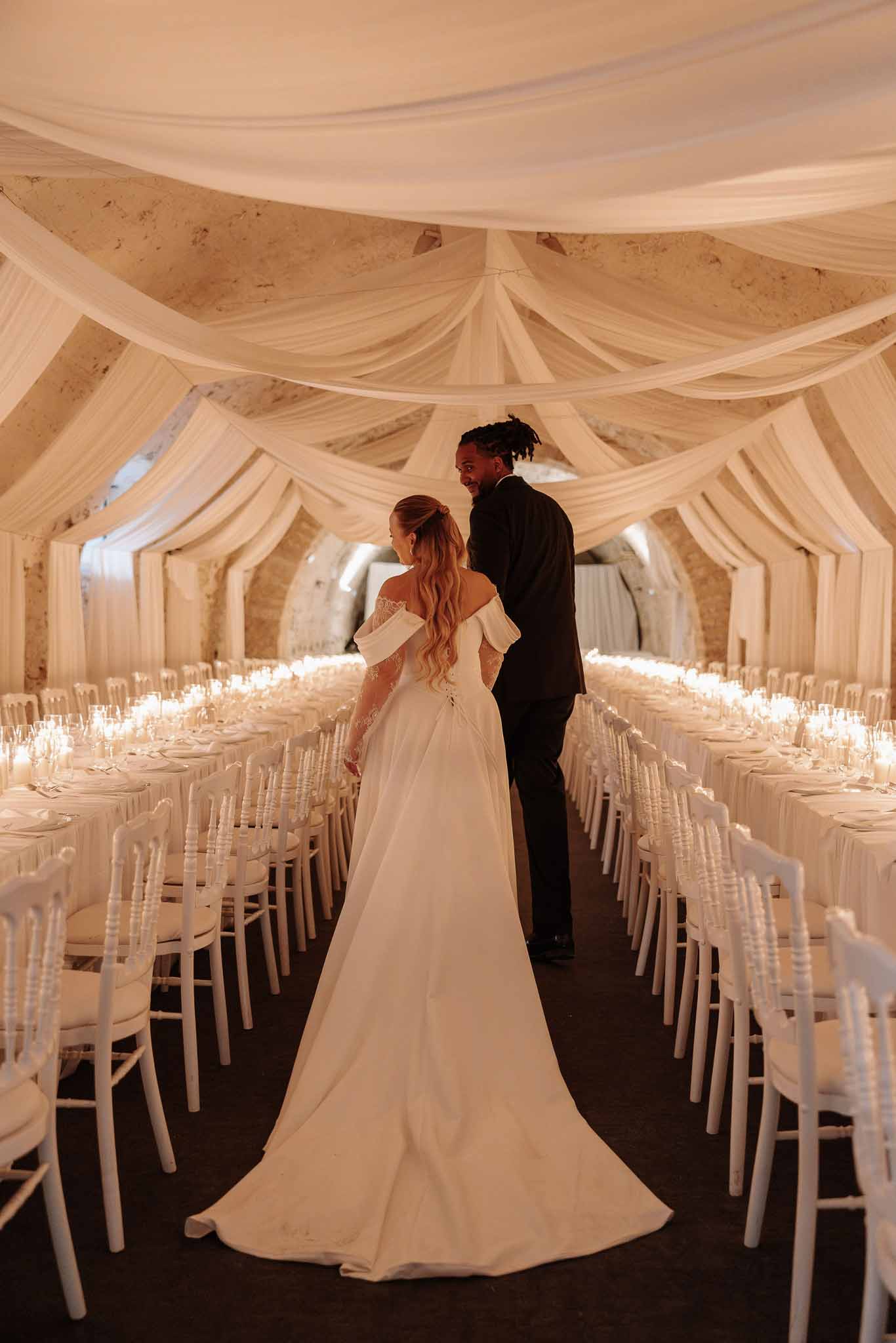 Couple entering draped reception hall with ivory ceiling fabric, candlelit banquet tables, and Chiavari chairs