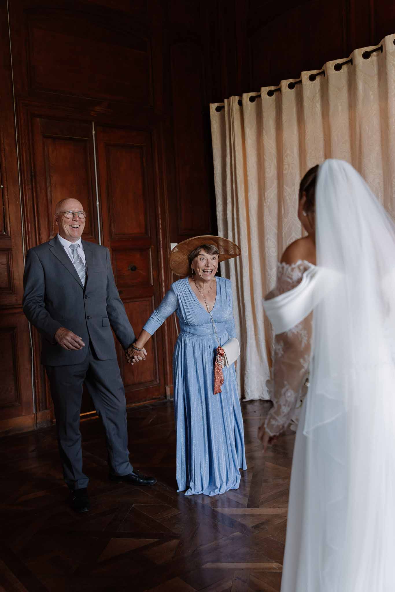 Bride in lace gown seen from behind as two guests react with joy during first look in chateau interior