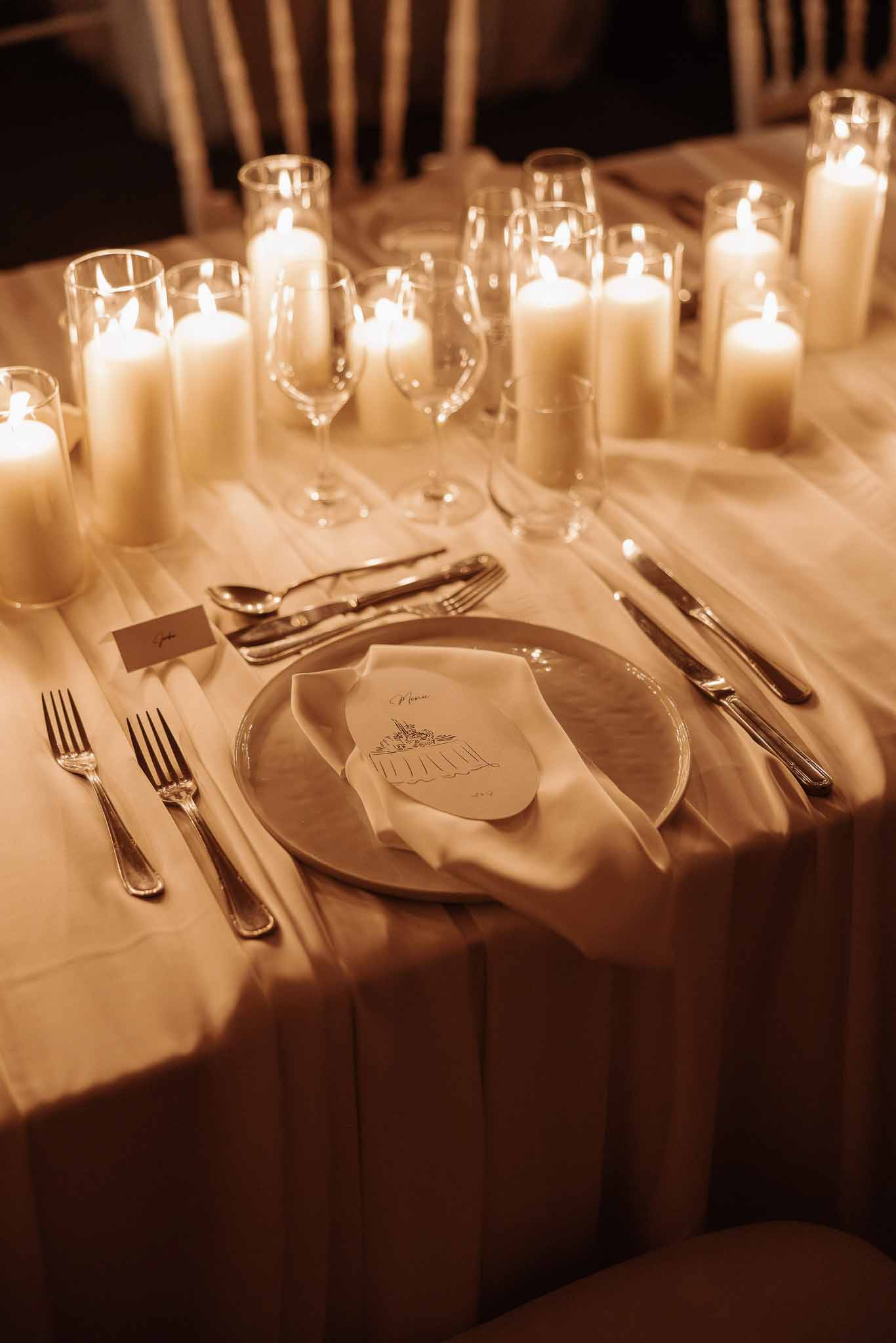 Candlelit place setting with illustrated venue menu card, silver cutlery, and ivory gathered cloth