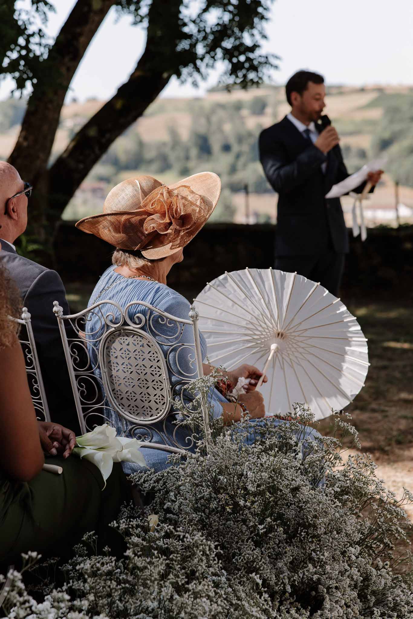 Guest in powder blue dress and caramel fascinator holding parasol beside white statice flowers at garden ceremony