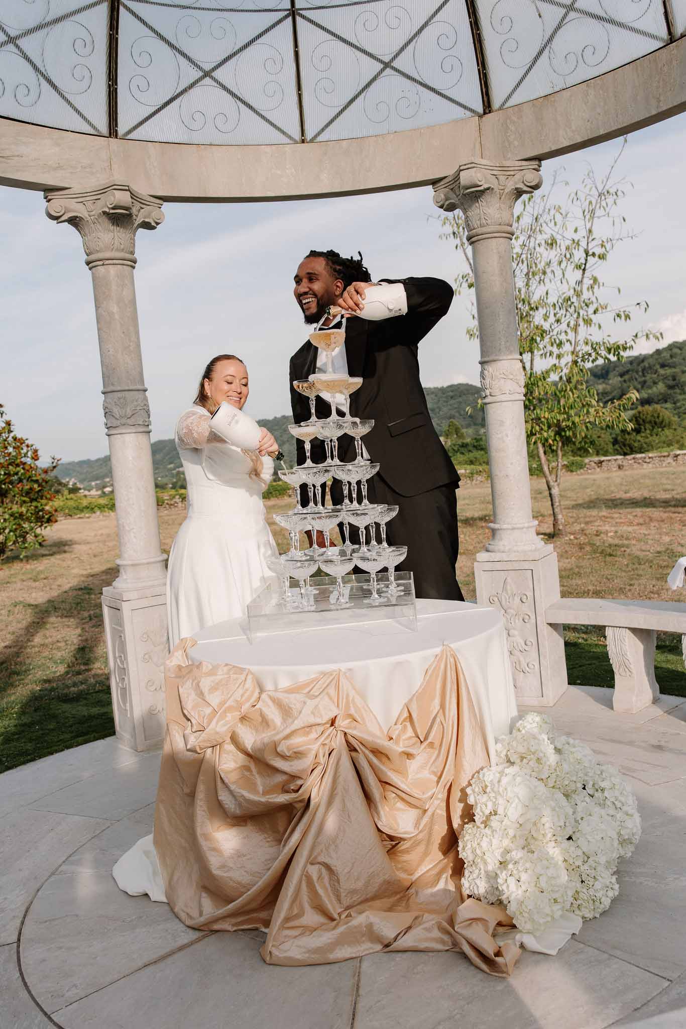 Bride and groom laughing while pouring champagne tower under stone gazebo with white hydrangea bouquets