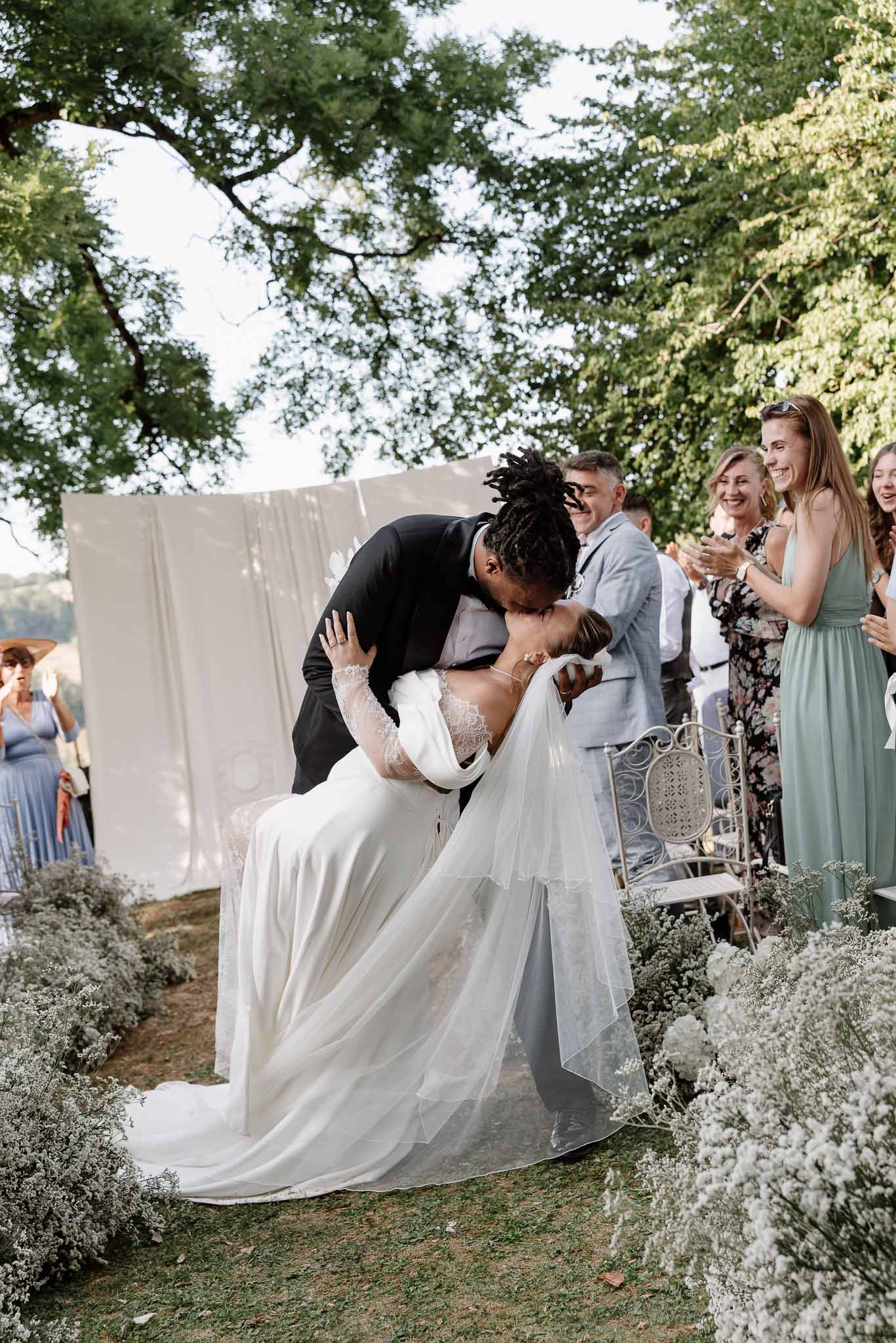 Bride and groom share dip kiss at outdoor ceremony under trees with baby's breath aisle decor and guests applauding
