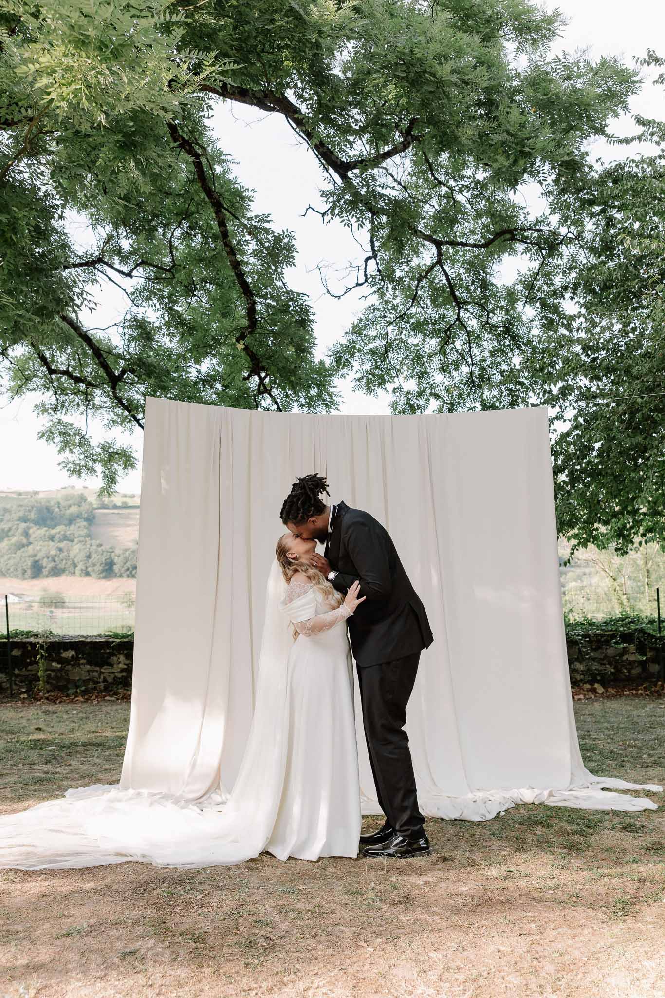 Couple kissing before cream draped backdrop between trees bride in off-shoulder lace gown with cathedral train groom in tu...