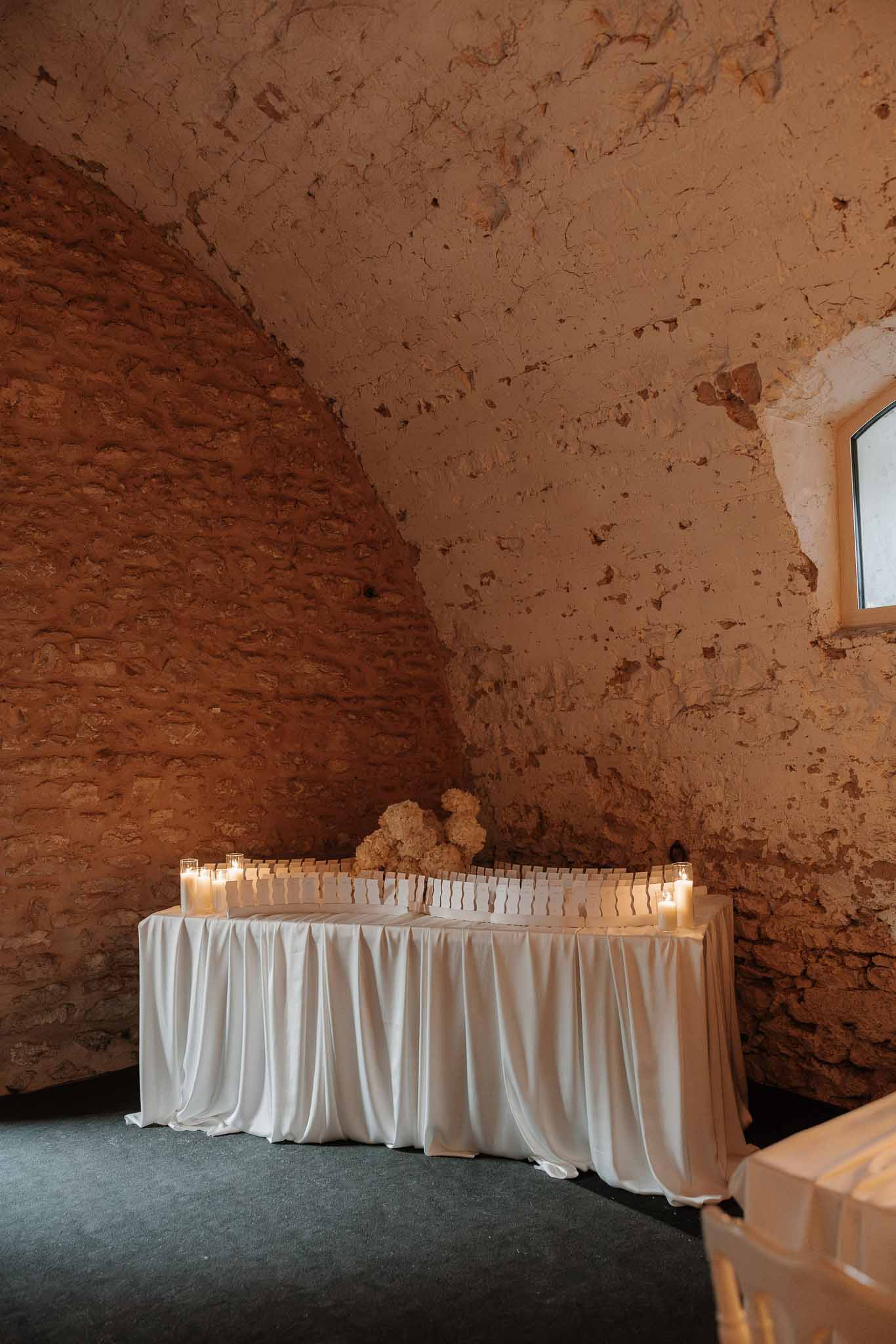 Escort card display table with white satin draping, pillar candles, and ivory floral arrangements in a vaulted stone room