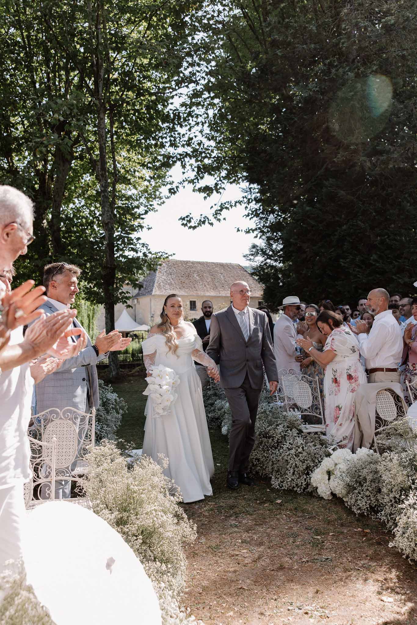 Bride walking down a garden aisle with her father past guests and baby's breath arrangements