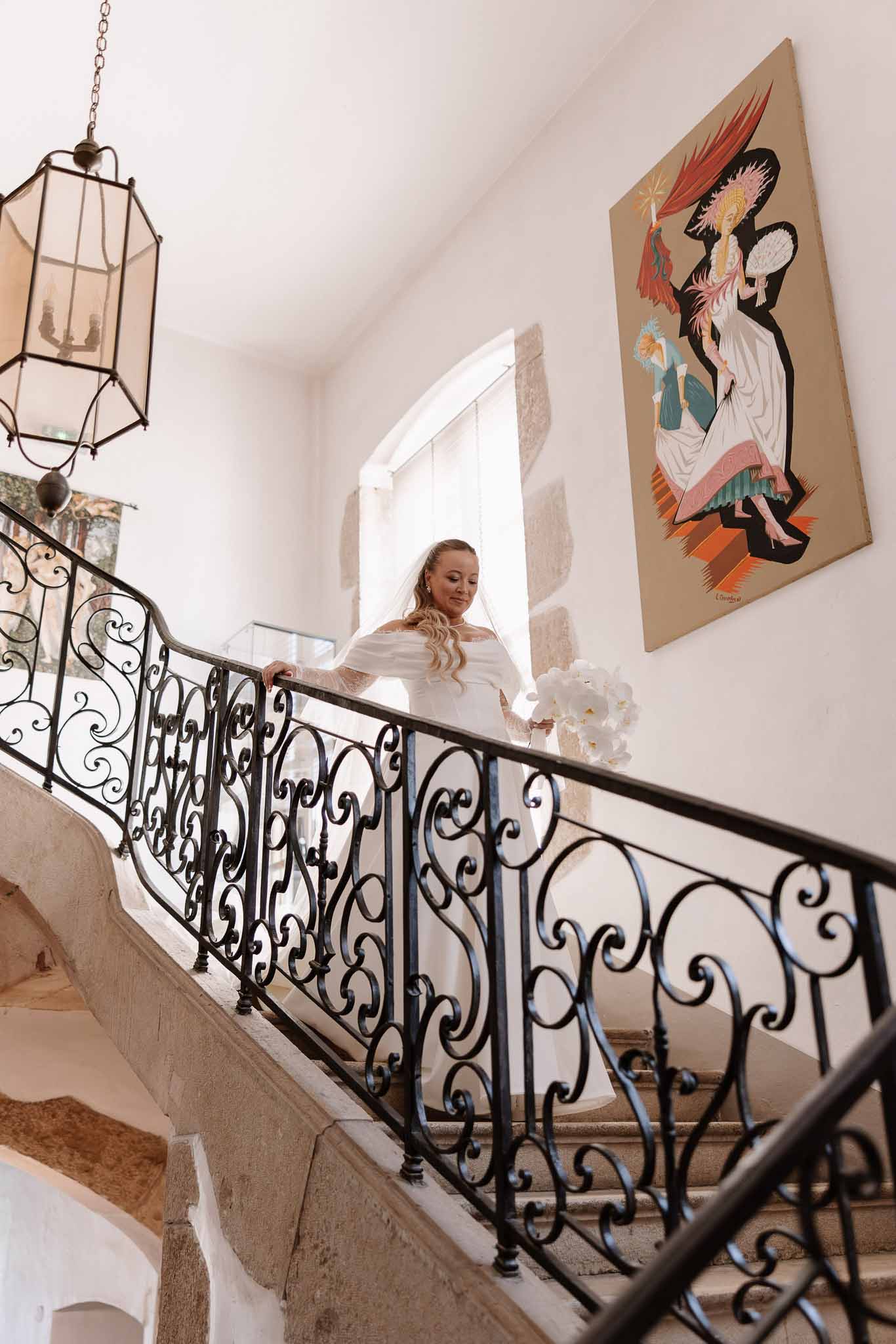 Bride descending wrought-iron staircase in off-shoulder gown with veil carrying white orchid bouquet past vintage lantern