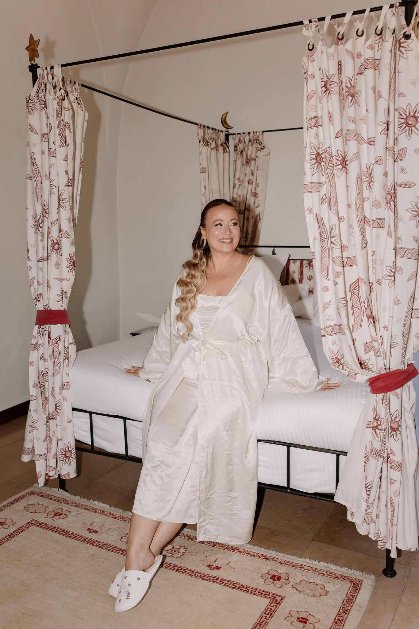 Bride in ivory satin robe on wrought-iron four-poster bed with red floral curtains and gold star finials