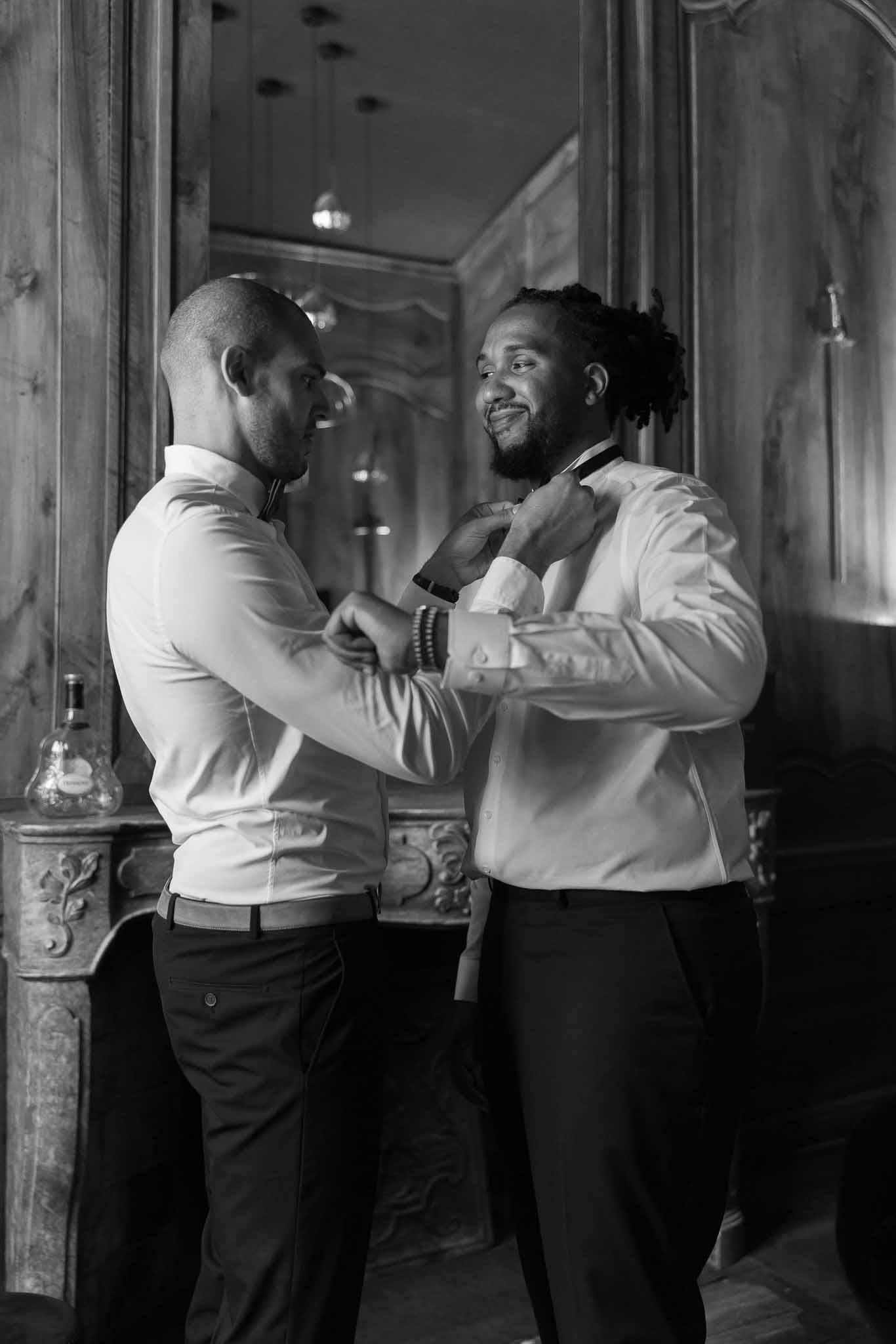 Black and white groomsman adjusting groom's bow tie before carved fireplace and ornate mirror in chateau