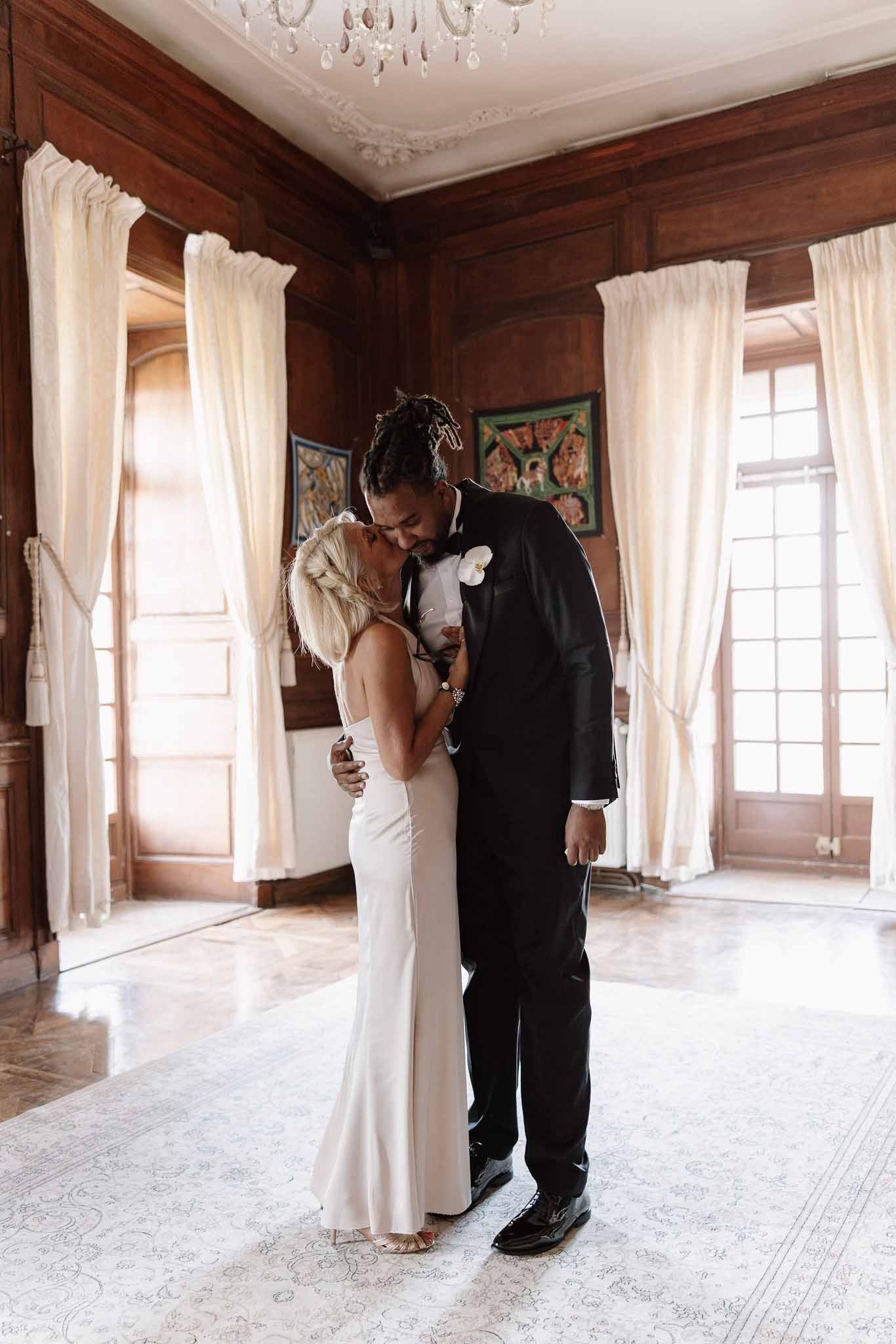 Bride in ivory slip gown and groom in black tuxedo kissing in chateau room with crystal chandelier and French windows