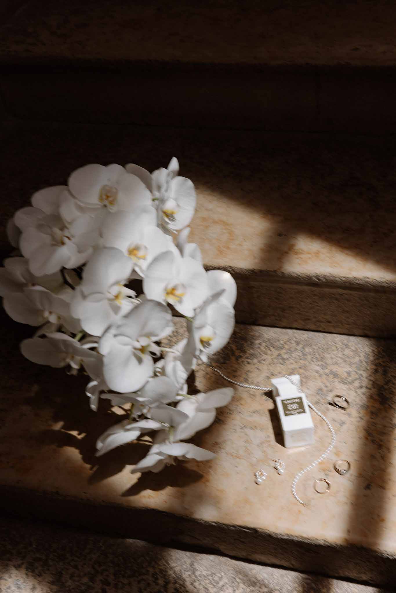 White orchid bouquet, Tom Ford perfume, pearl bracelet, and gold bands arranged on sunlit stone steps
