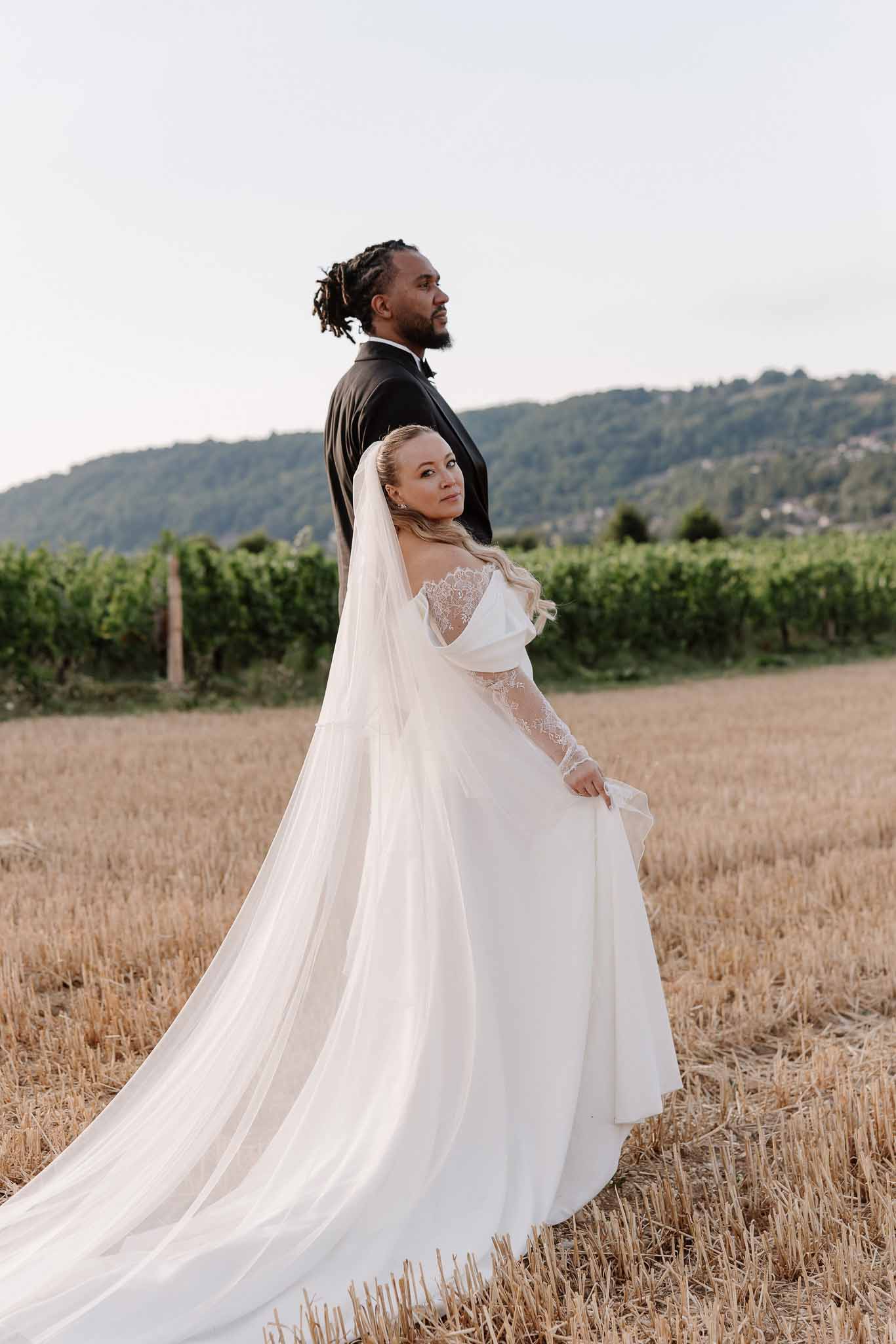 Groom in black tuxedo and bride in lace-sleeve gown with cathedral veil in golden hour harvested wheat field