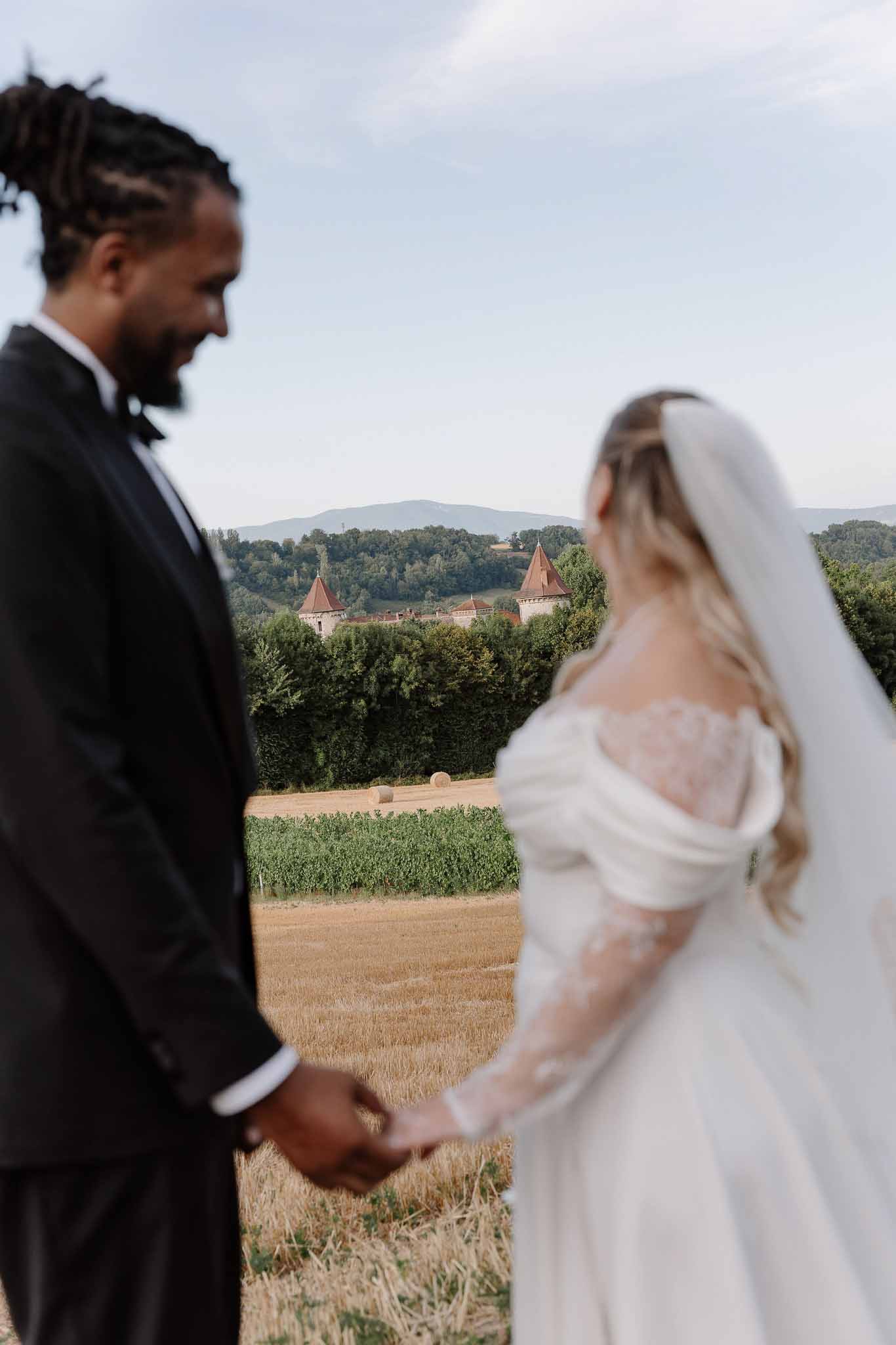 Bride and groom holding hands facing a French chateau with conical towers beyond harvested wheat fields