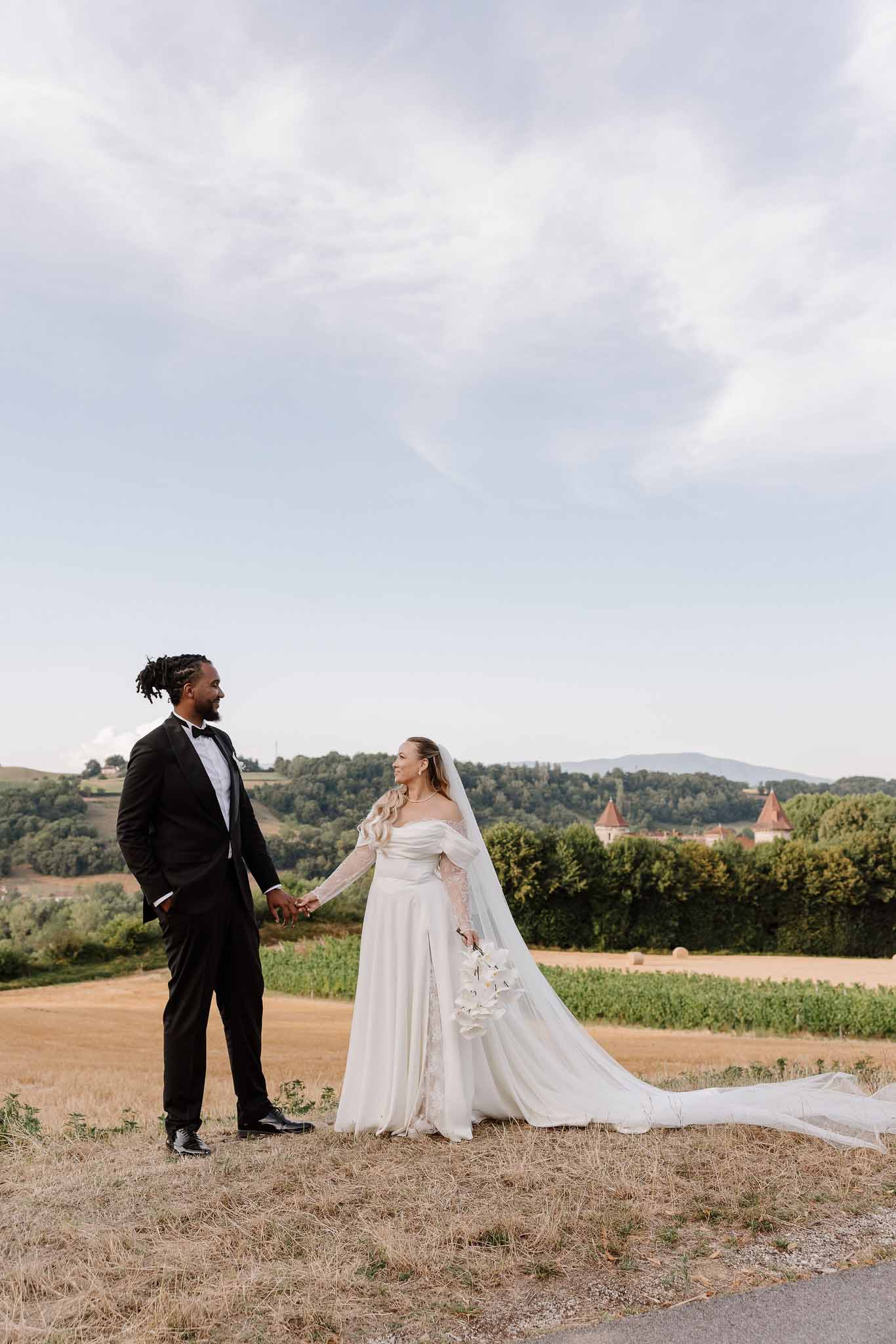 Bride in off-shoulder lace gown with cathedral veil and groom in black tuxedo holding hands in French countryside