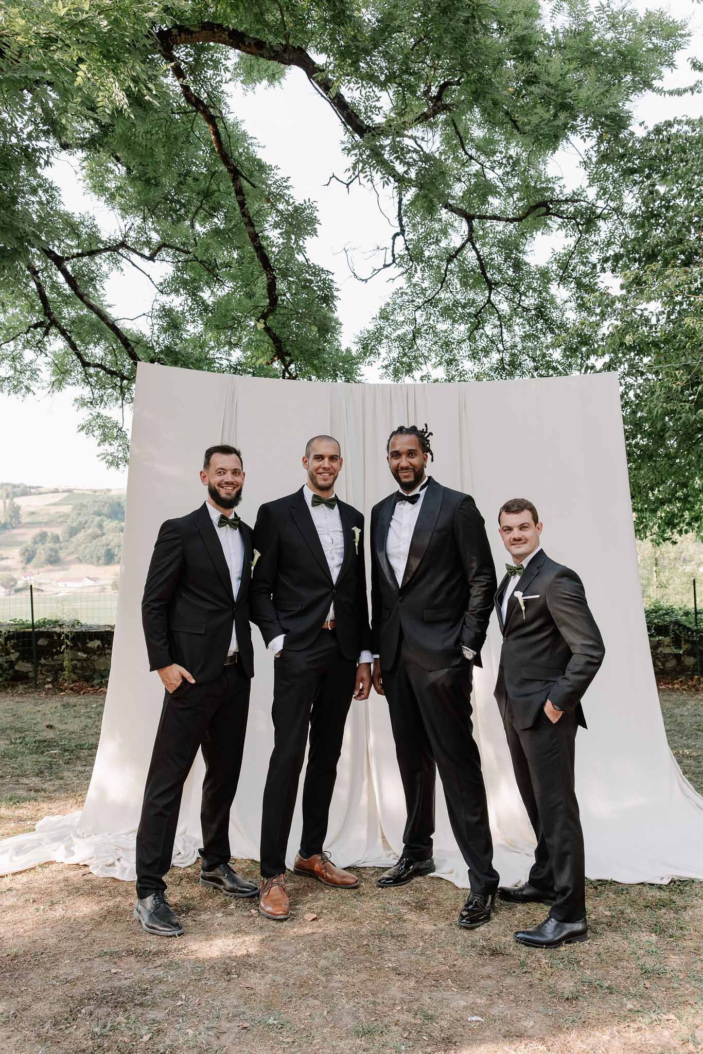 Groom and three groomsmen in black suits with olive green bow ties posing before ivory backdrop outdoors