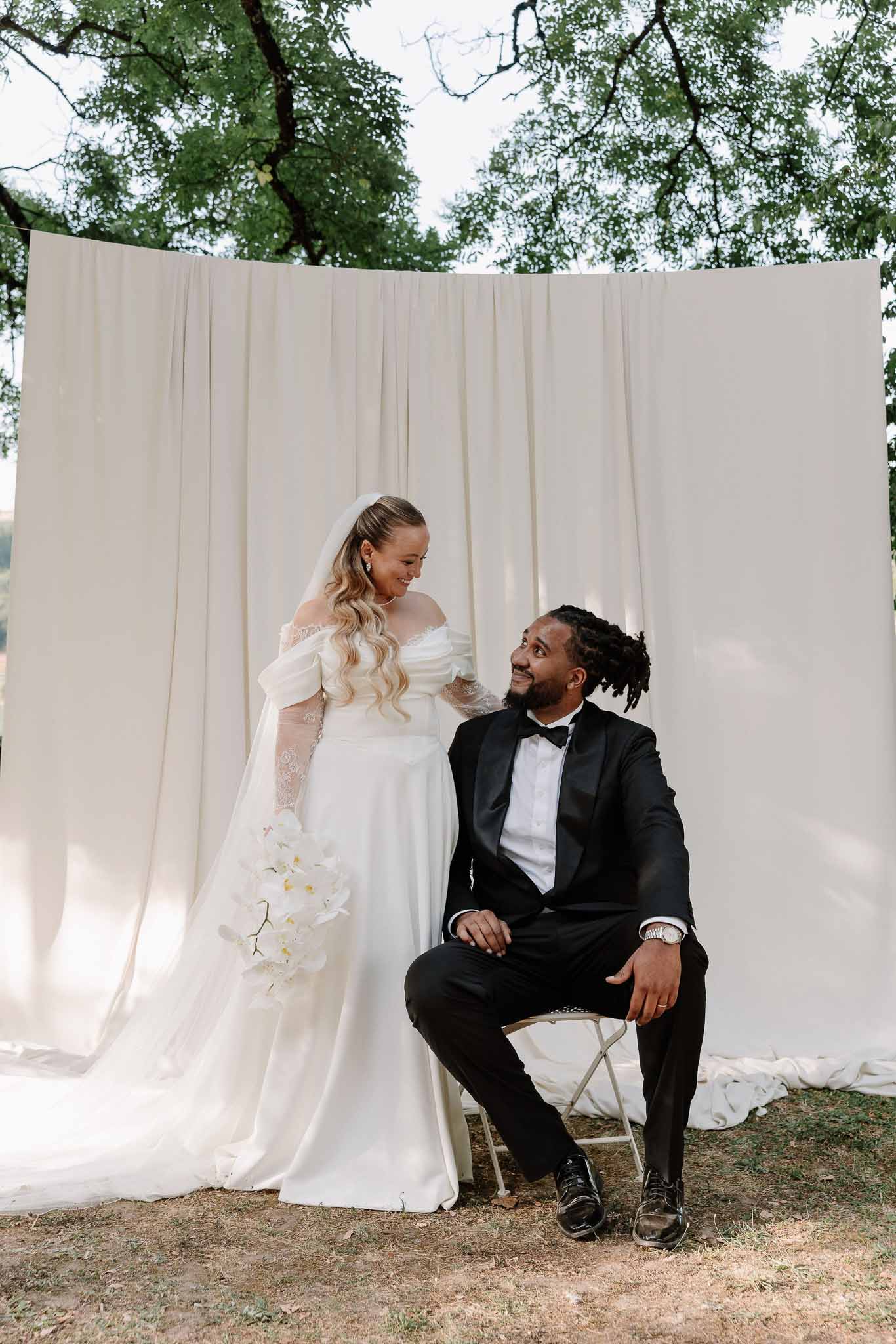 Bride in puff-sleeve gown with orchid cascade and groom in tuxedo seated against ivory drape backdrop