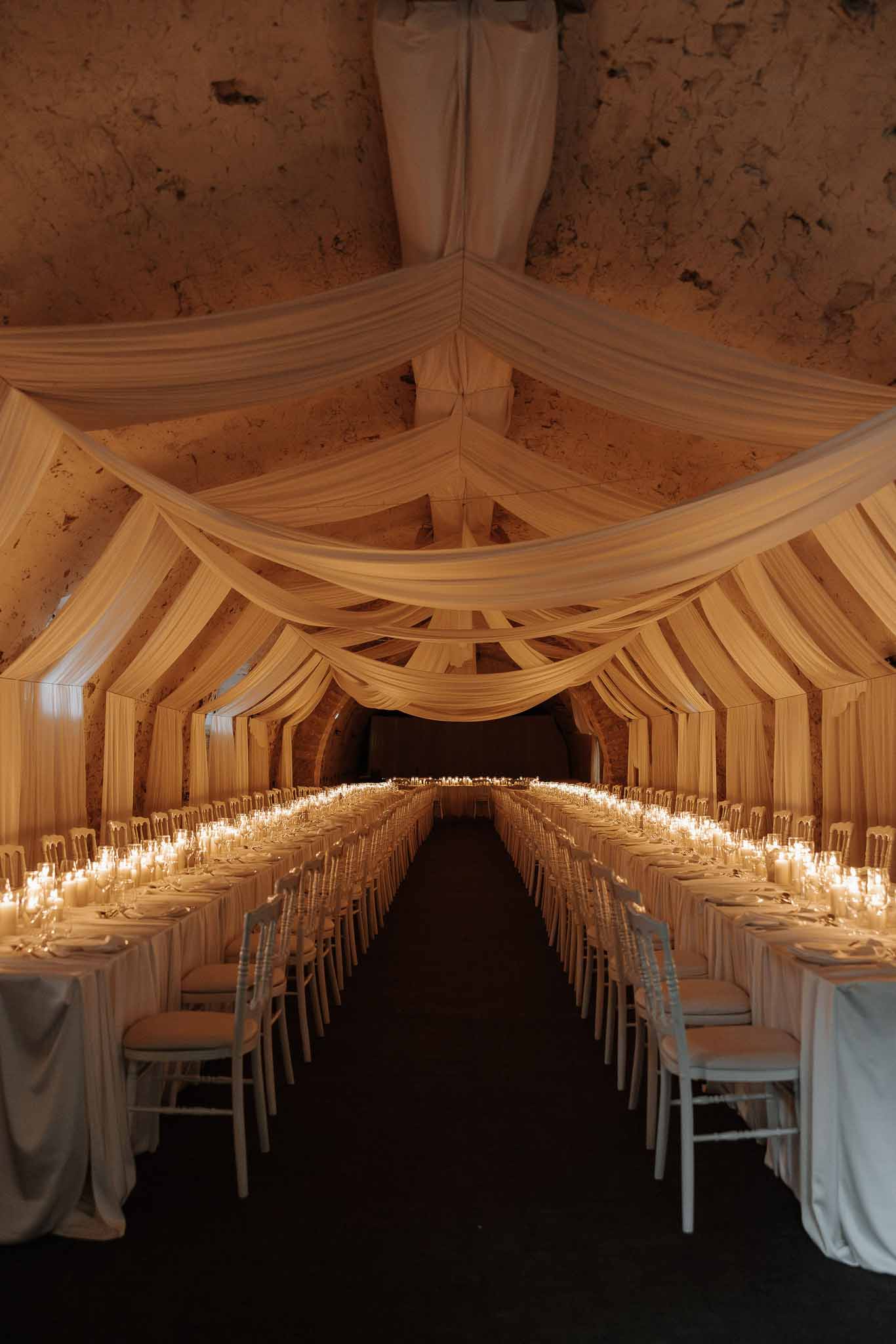 Two long tables in ivory-draped vaulted cave with hundreds of candles and white chiavari chairs