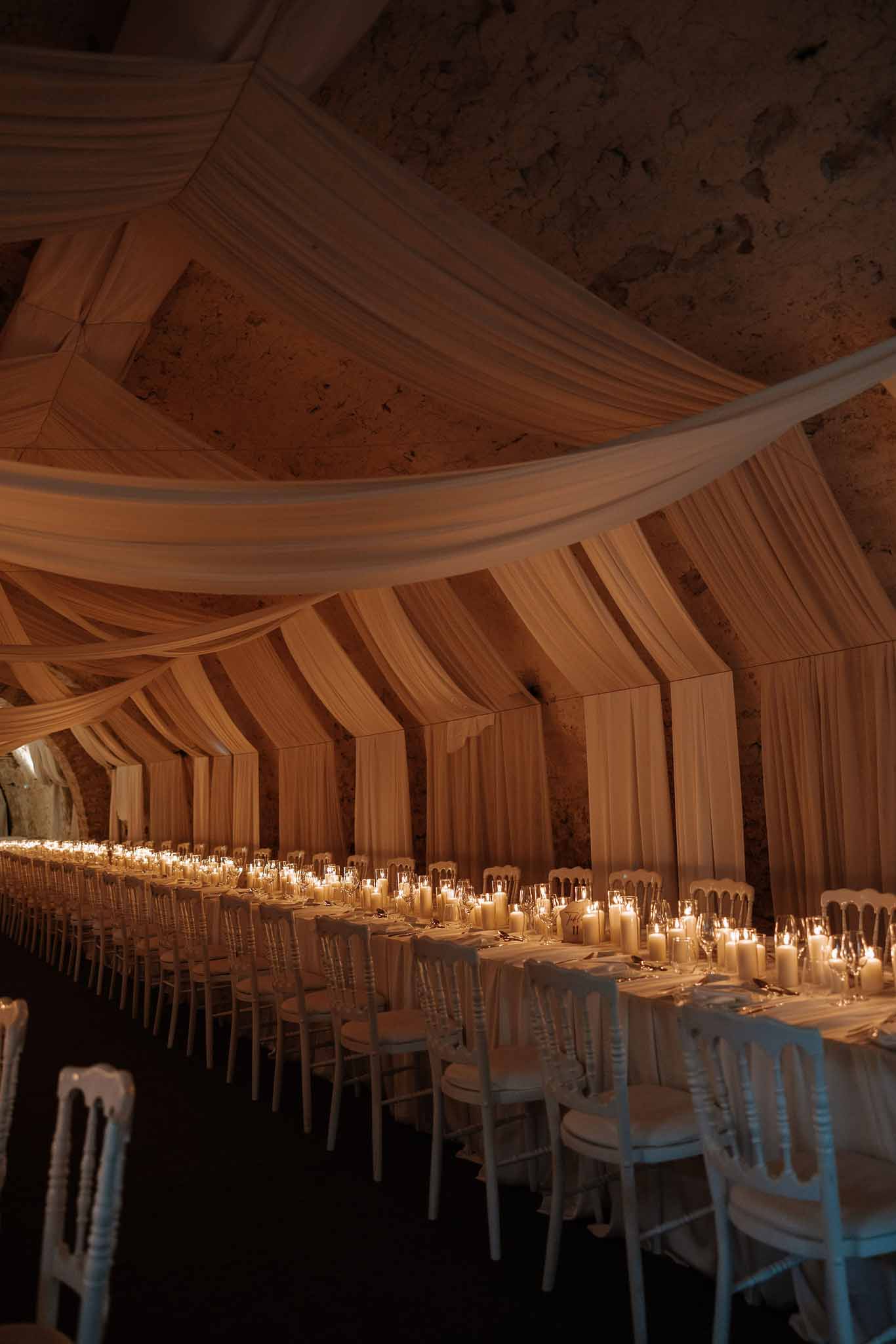Wedding reception setup inside a vaulted stone building with ivory draping and long tables lit by pillar candles