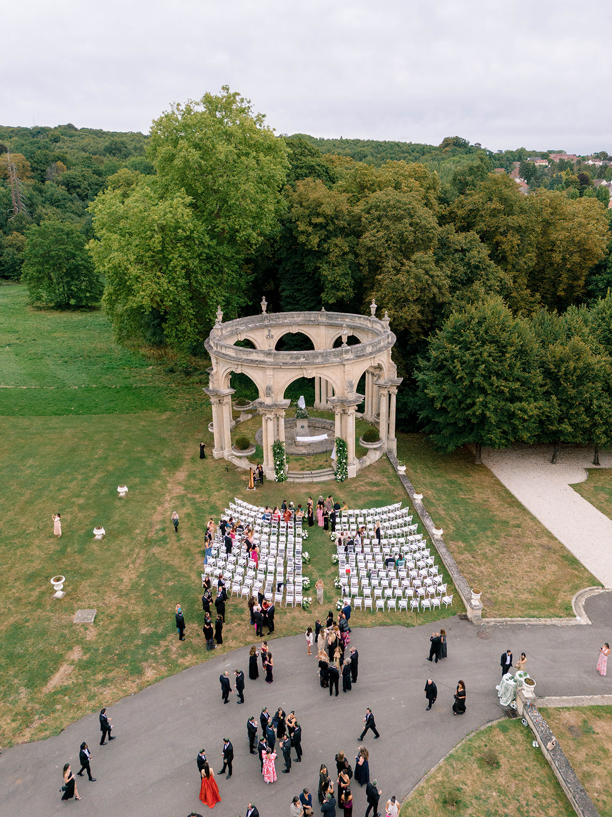 Aerial view of outdoor wedding ceremony at neoclassical stone rotunda with white chairs and guests on chateau grounds