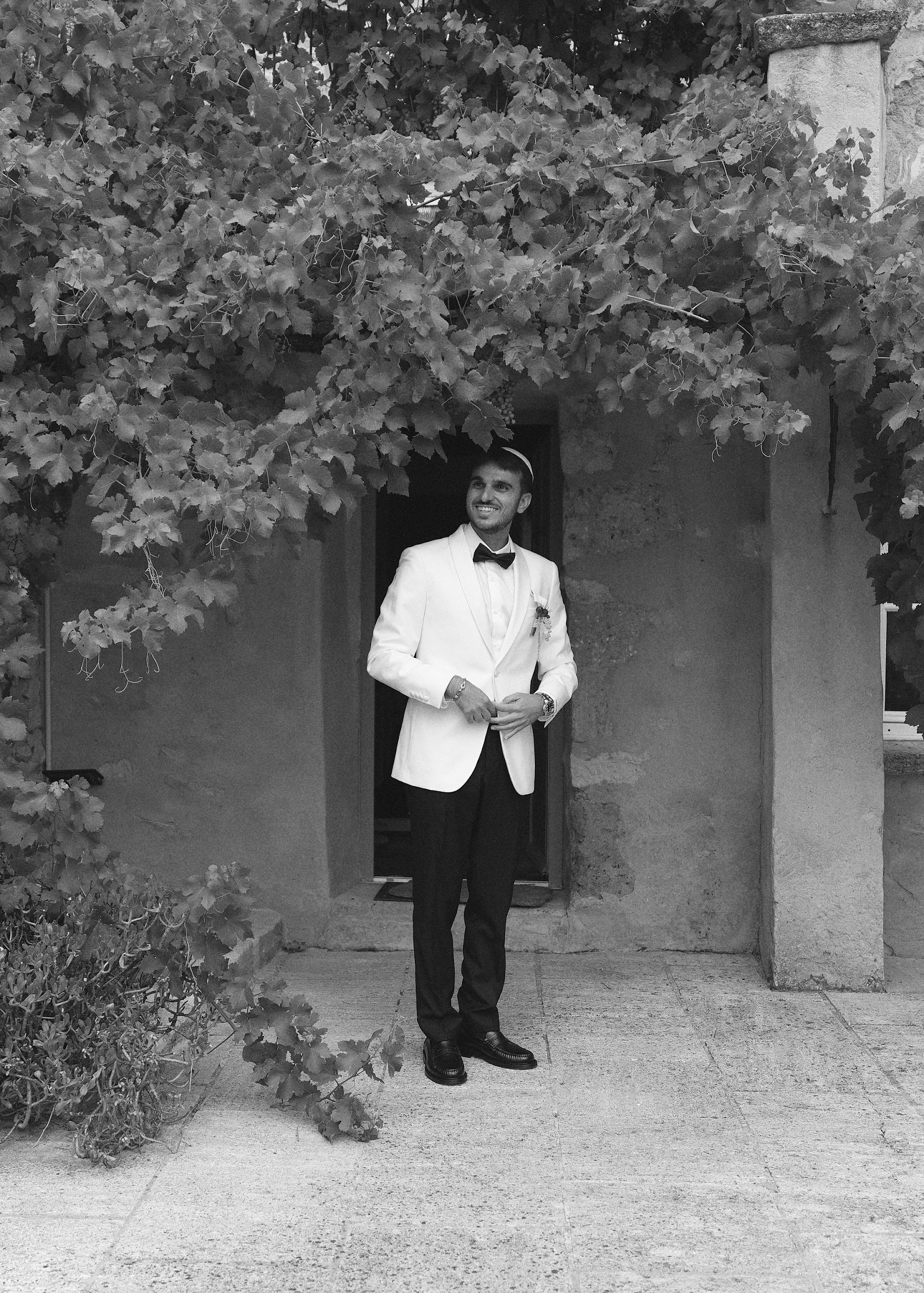 Black and white groom in white dinner jacket with kippah smiling in vine-draped stone doorway