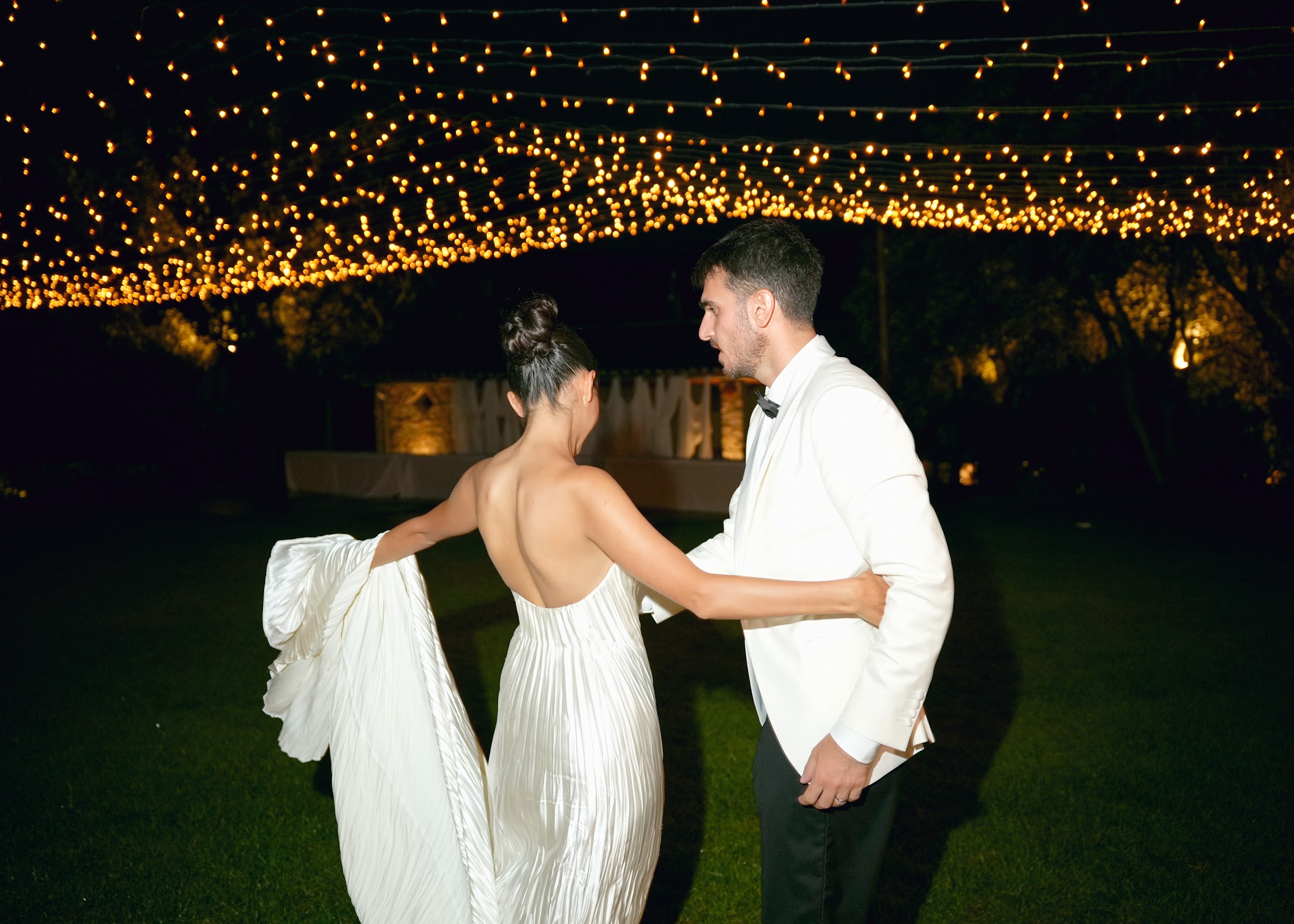 Bride and groom walking under fairy light canopy at night with dramatic draped train