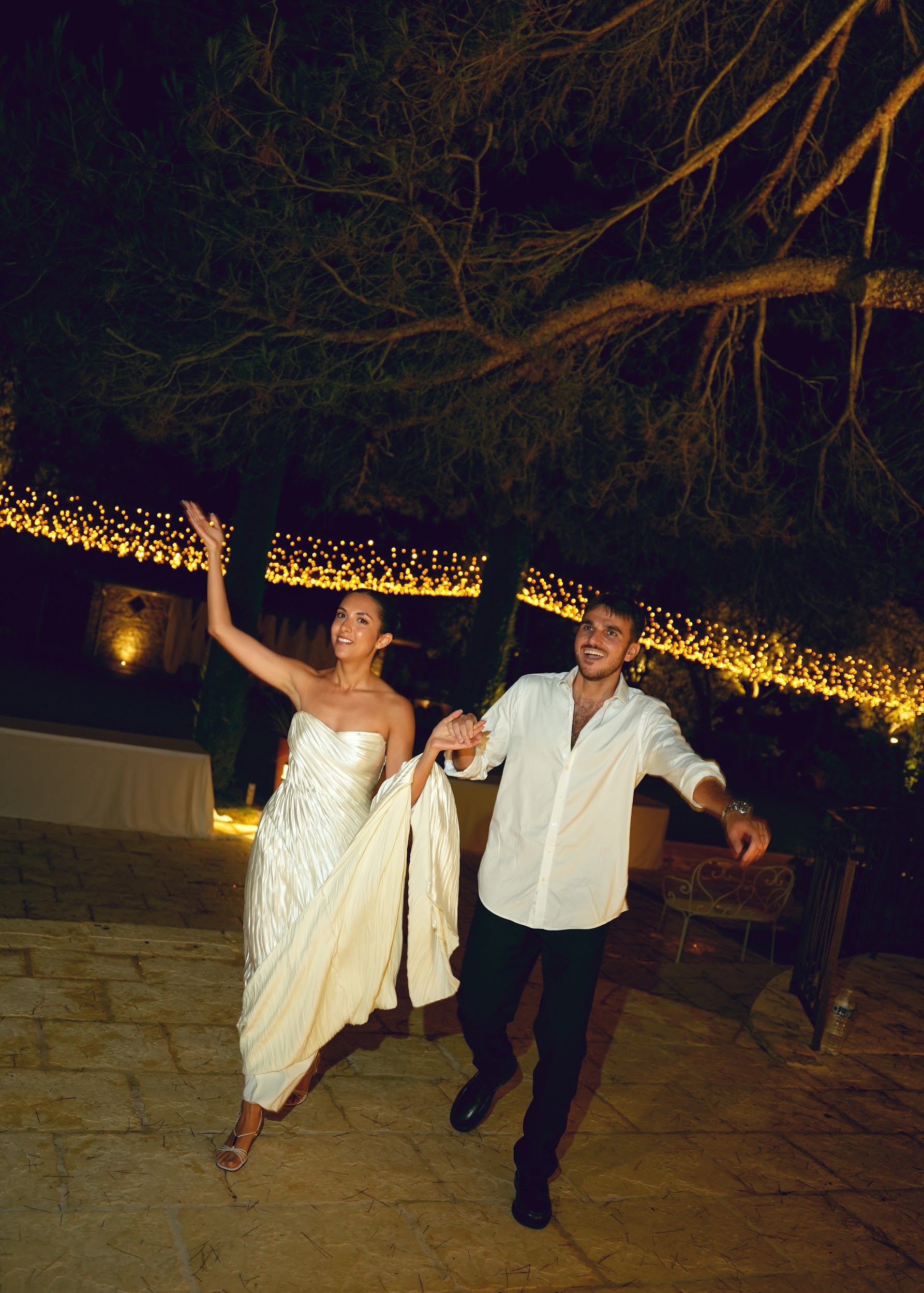 Couple dancing on stone terrace at night with golden fairy lights bride in ivory pleated satin gown