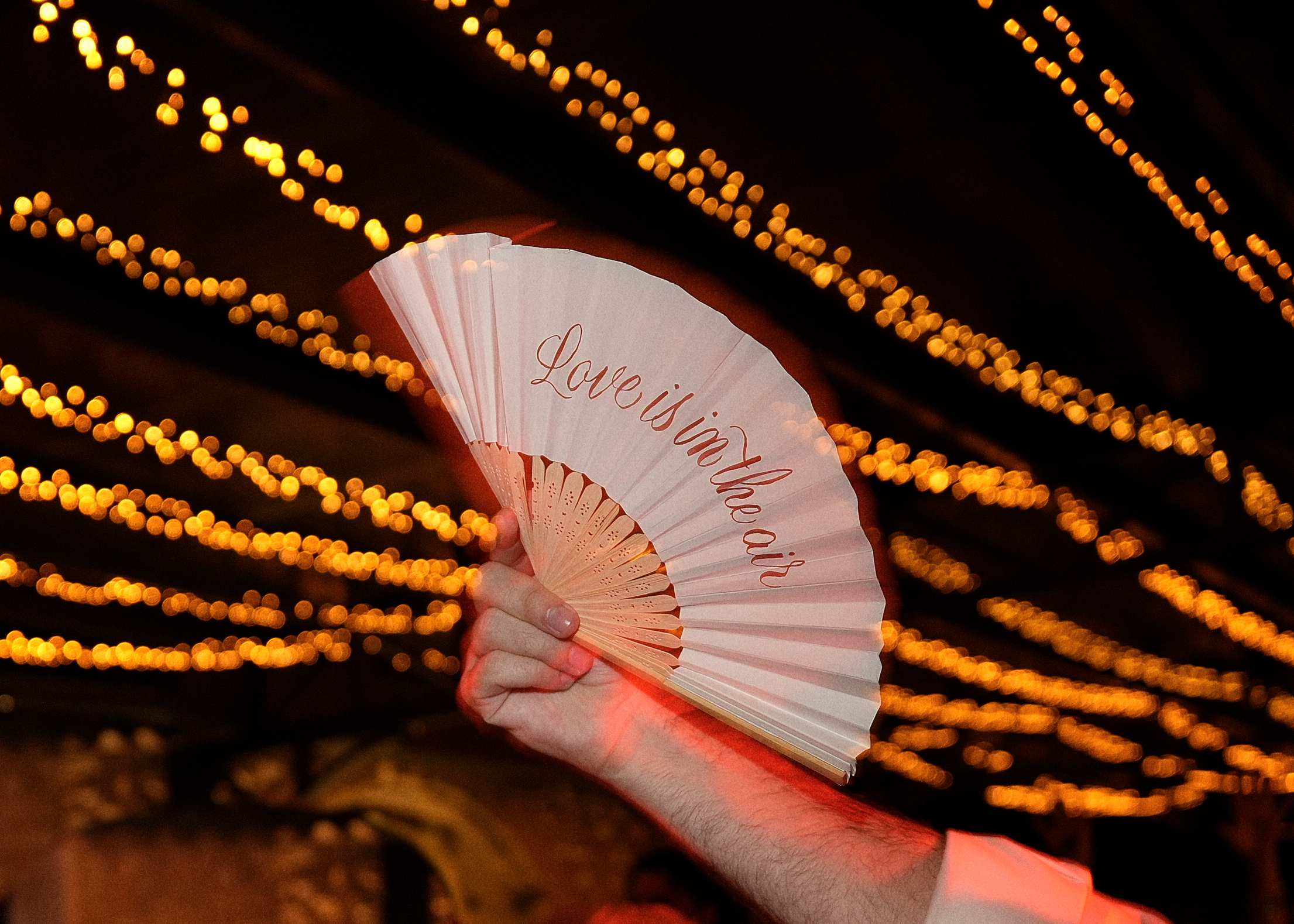 White paper fan reading Love is in the air held against golden fairy light canopy at evening reception
