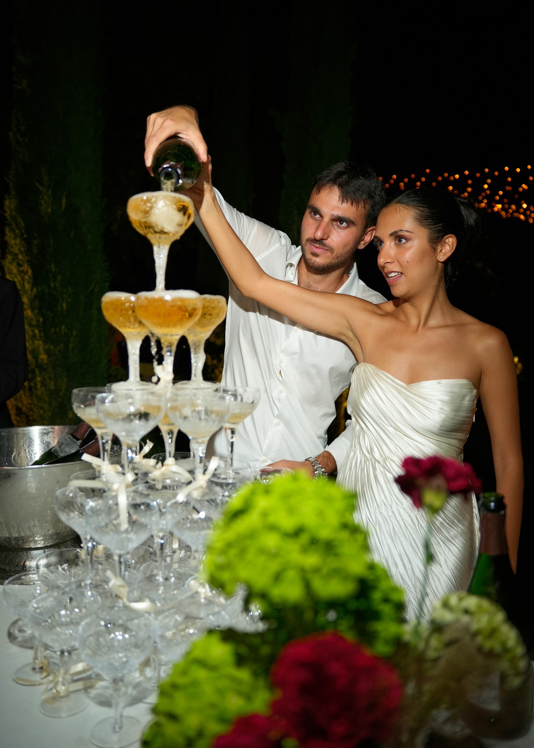 Bride and groom pouring champagne into a coupe glass tower during evening outdoor reception