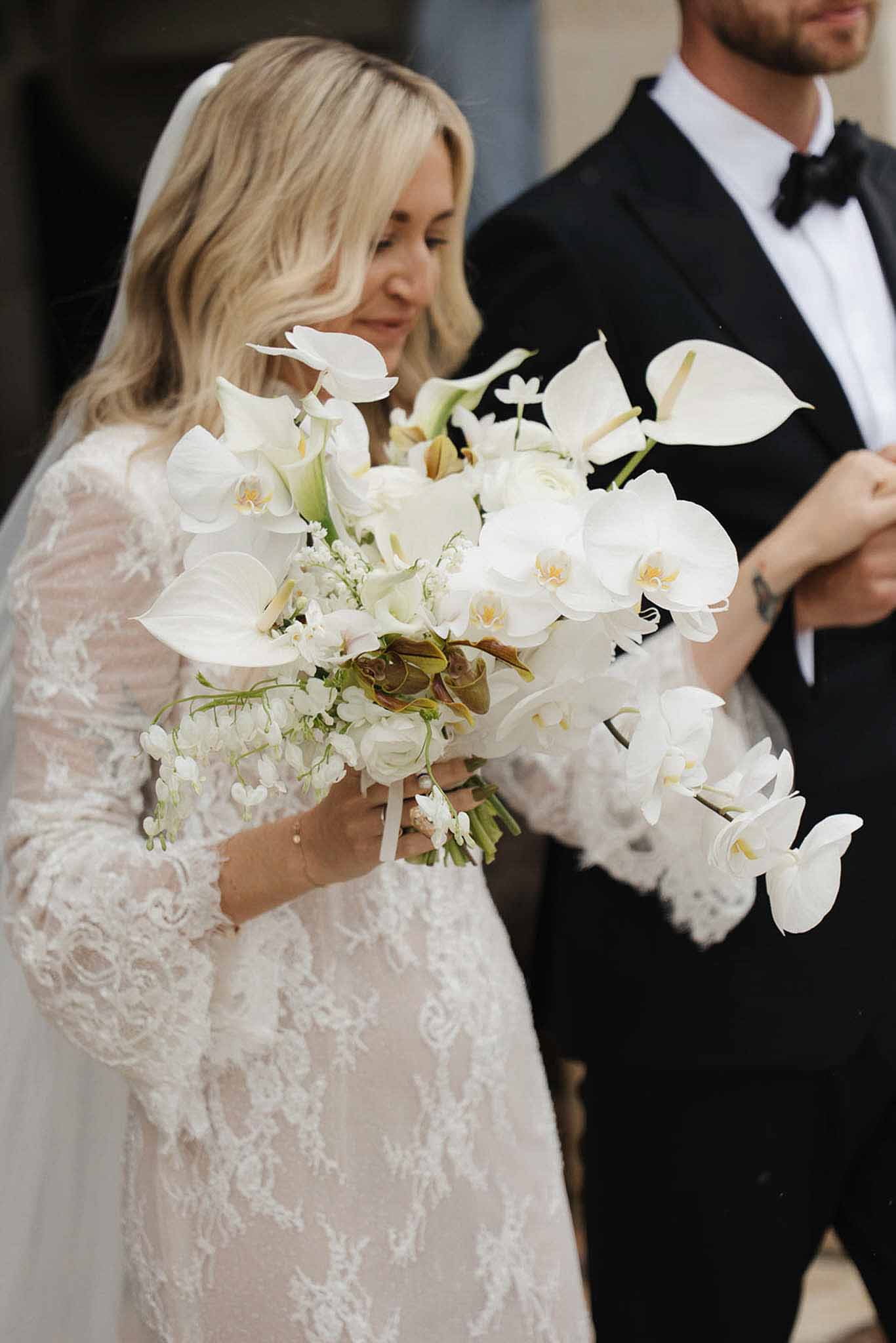 Bride in full-lace gown with cascading white orchid and calla lily bouquet beside groom in tuxedo