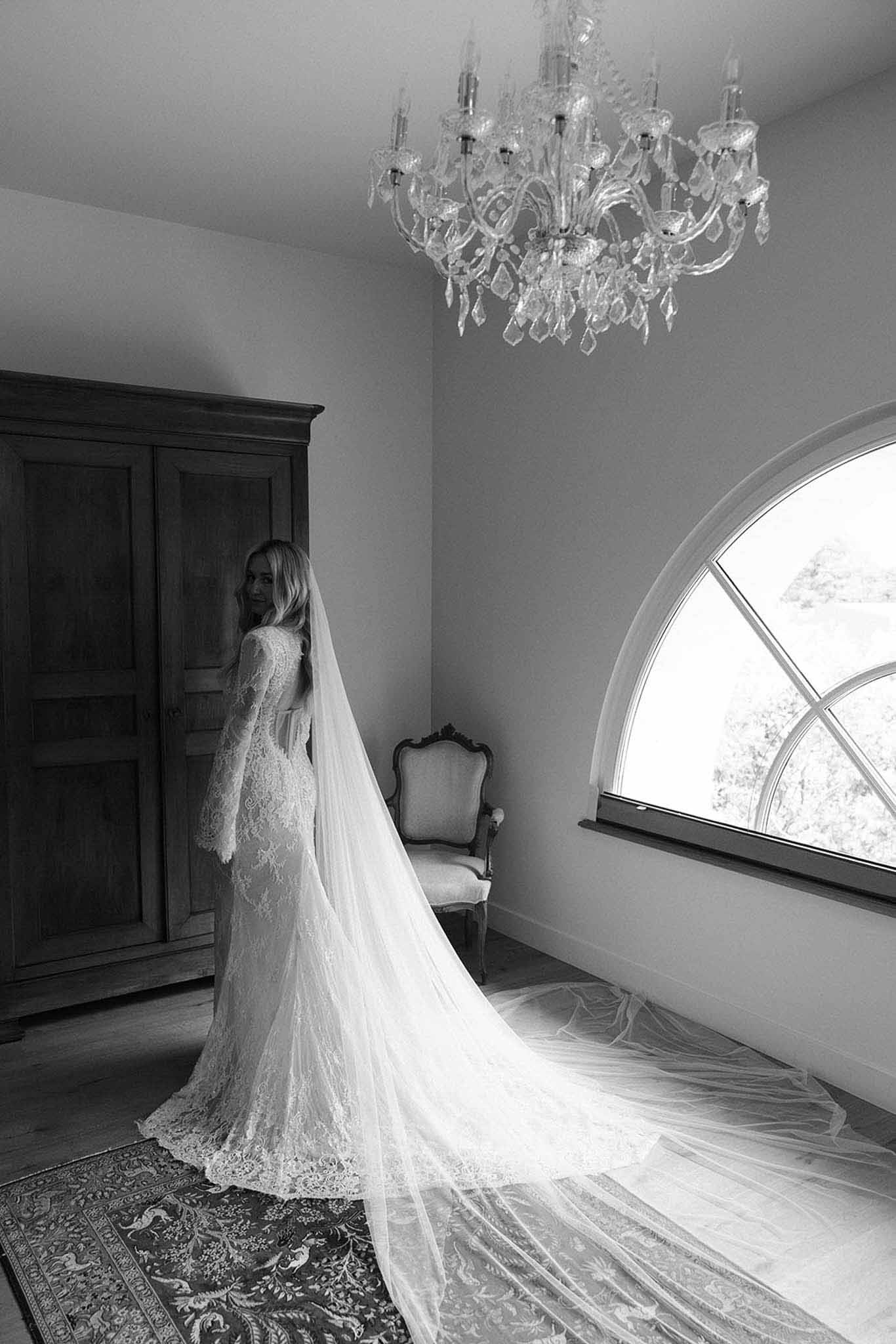 Black-and-white portrait of bride in long-sleeve lace gown with cathedral veil in chateau room with chandelier