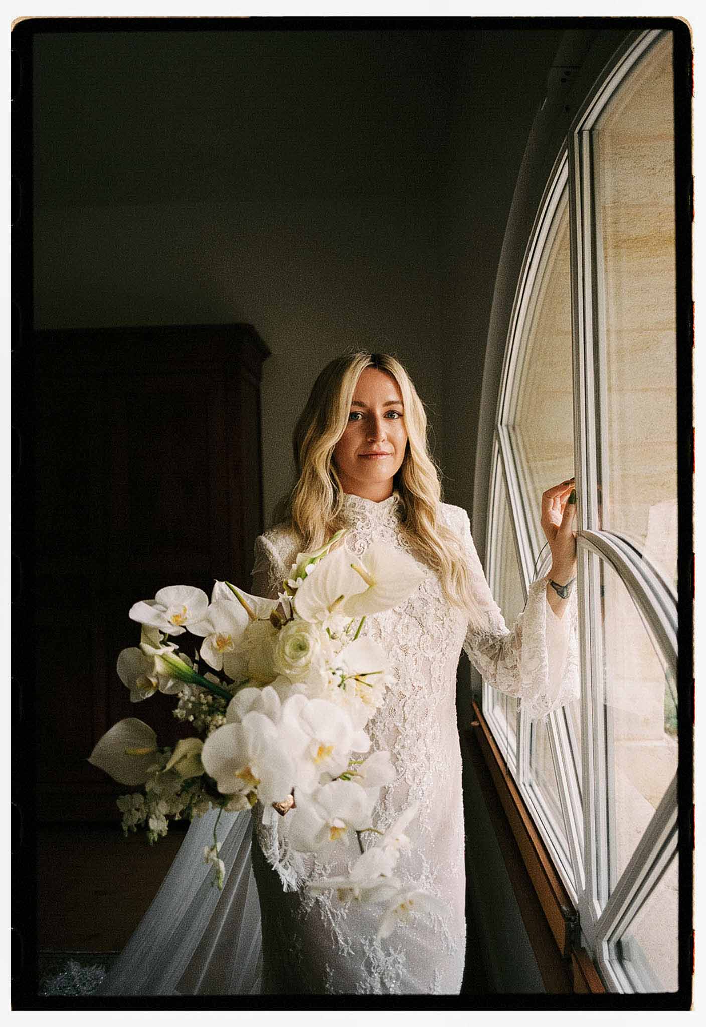 Bride in long-sleeve ivory lace gown holding cascading white orchid bouquet by arched window in shadow