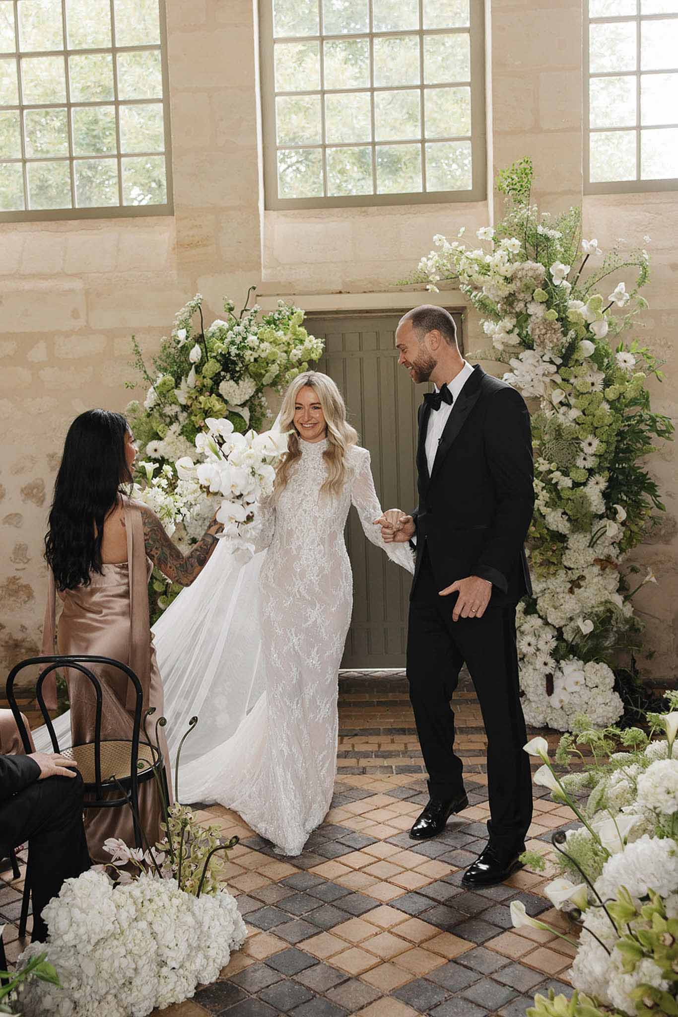 Bride and groom holding hands during ceremony in stone chapel with white orchid and hydrangea floral columns
