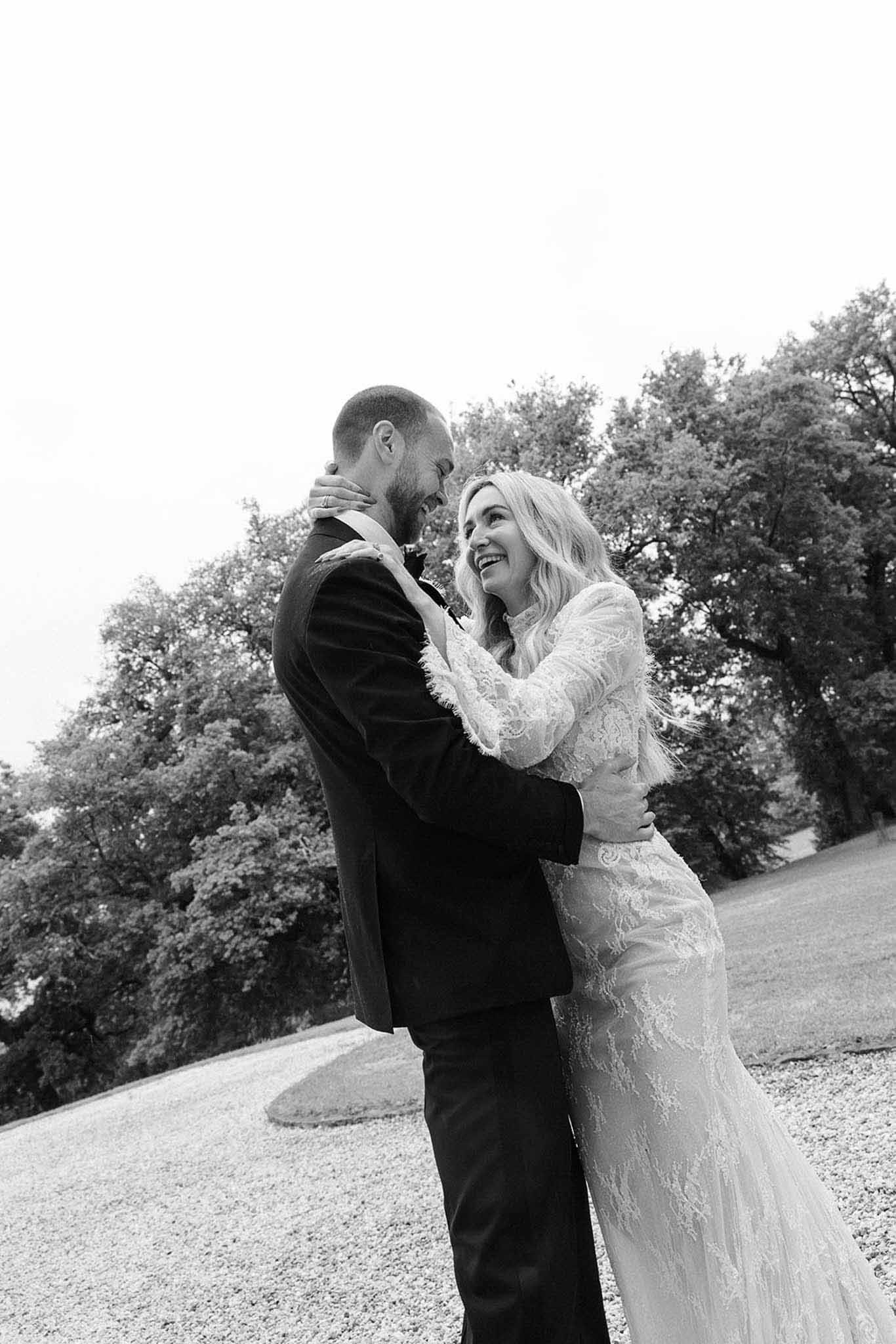 Bride in long-sleeve lace gown laughing with groom on estate gravel drive in soft B&W