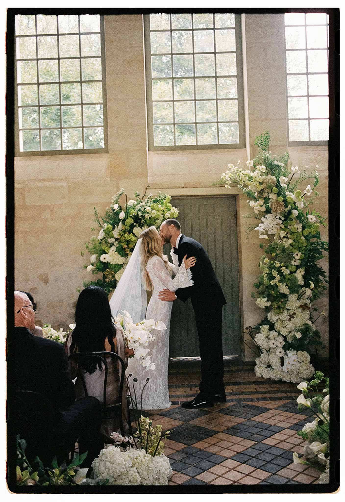 Bride and groom share first kiss at altar flanked by white floral installations in stone chapel