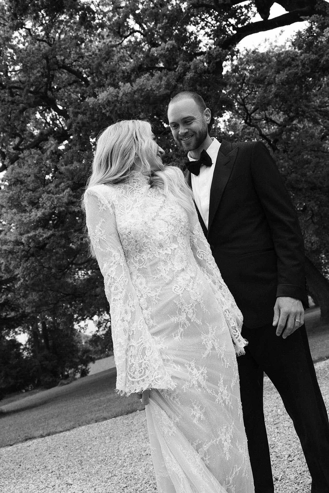 Black-and-white portrait of bride in long-sleeve lace gown and groom in tuxedo smiling on chateau grounds