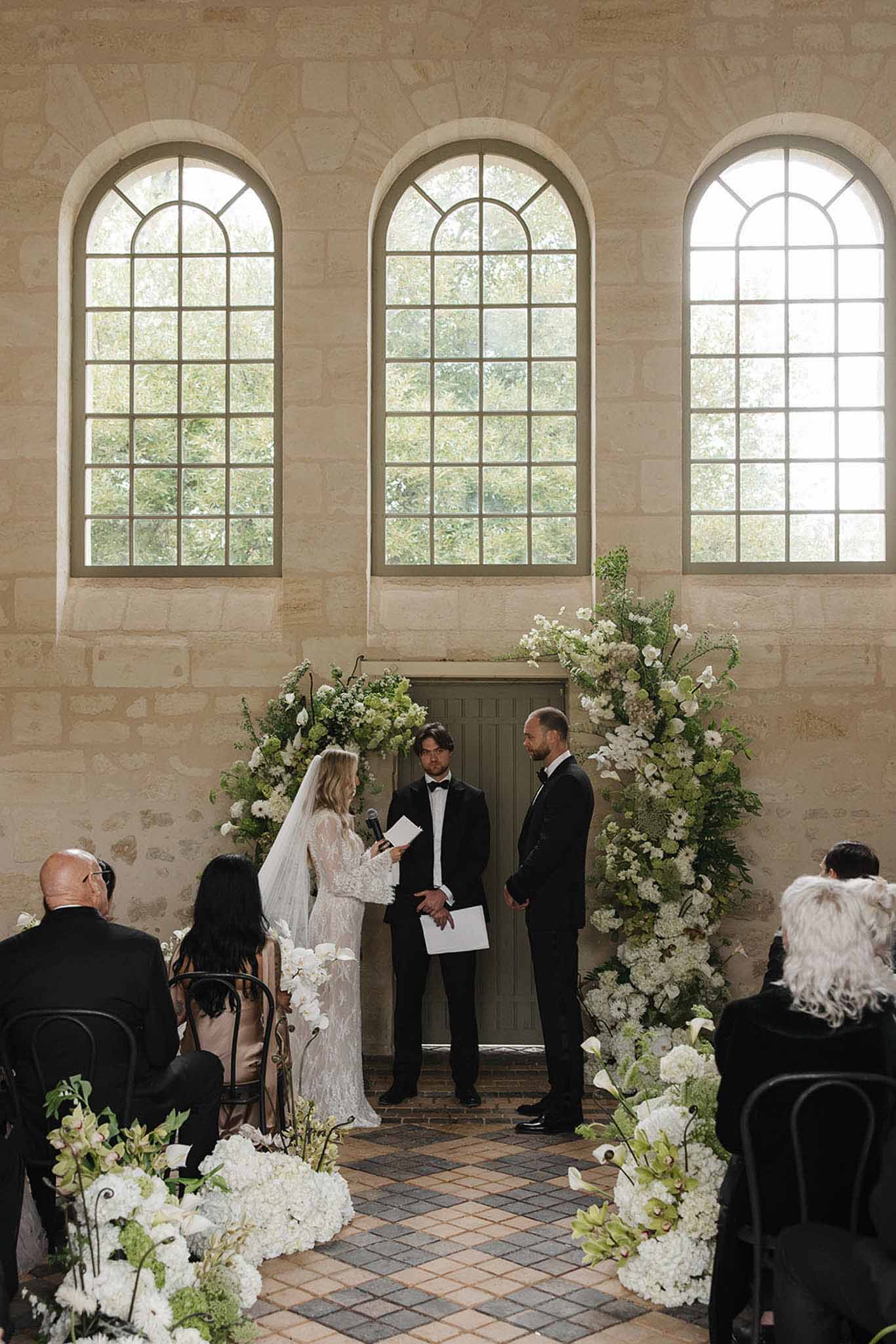 Bride reading vows to groom under a circular white floral arch in a stone chapel with arched windows
