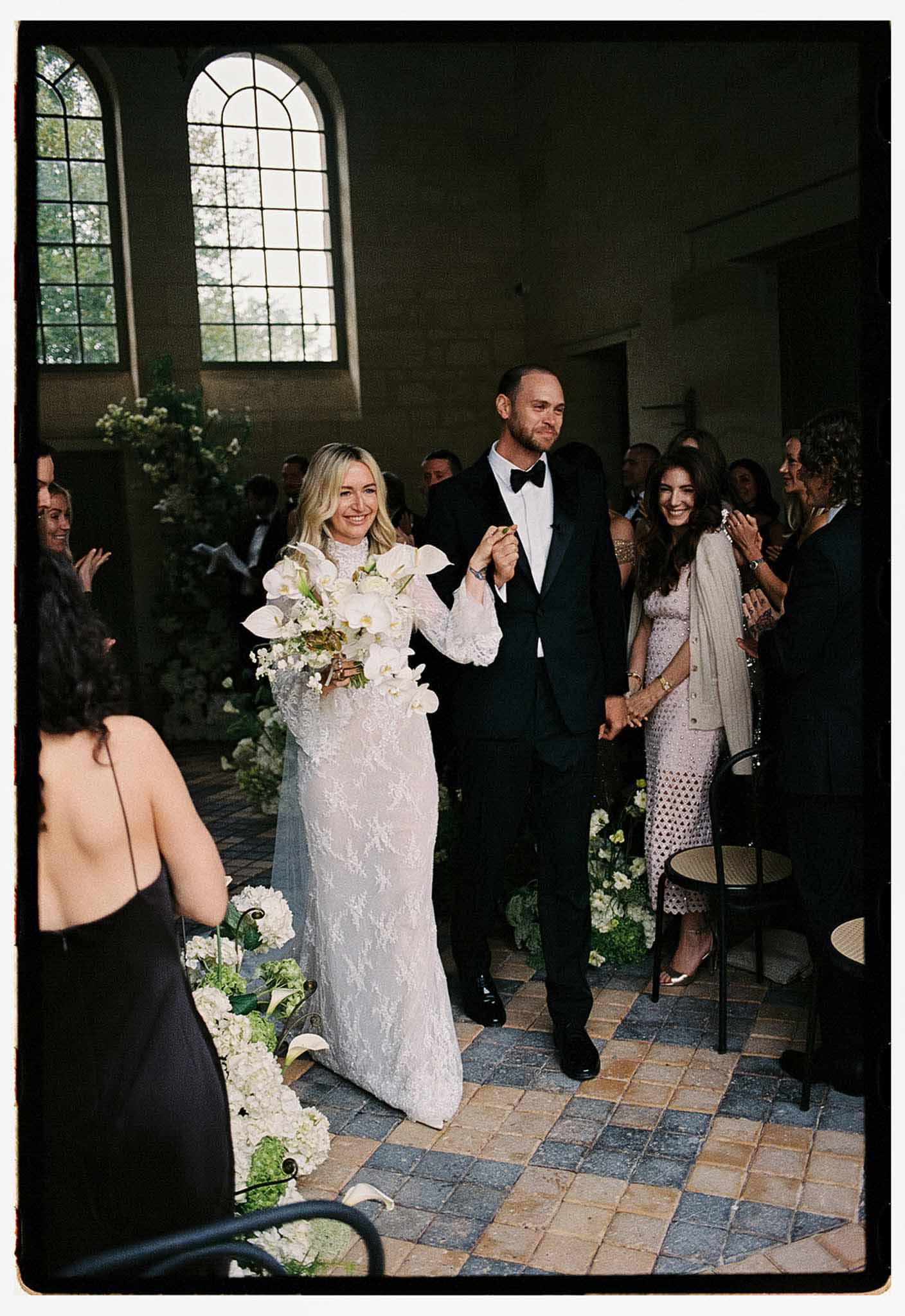 Couple walking aisle past white orchid and hydrangea arrangements in arched indoor ceremony space