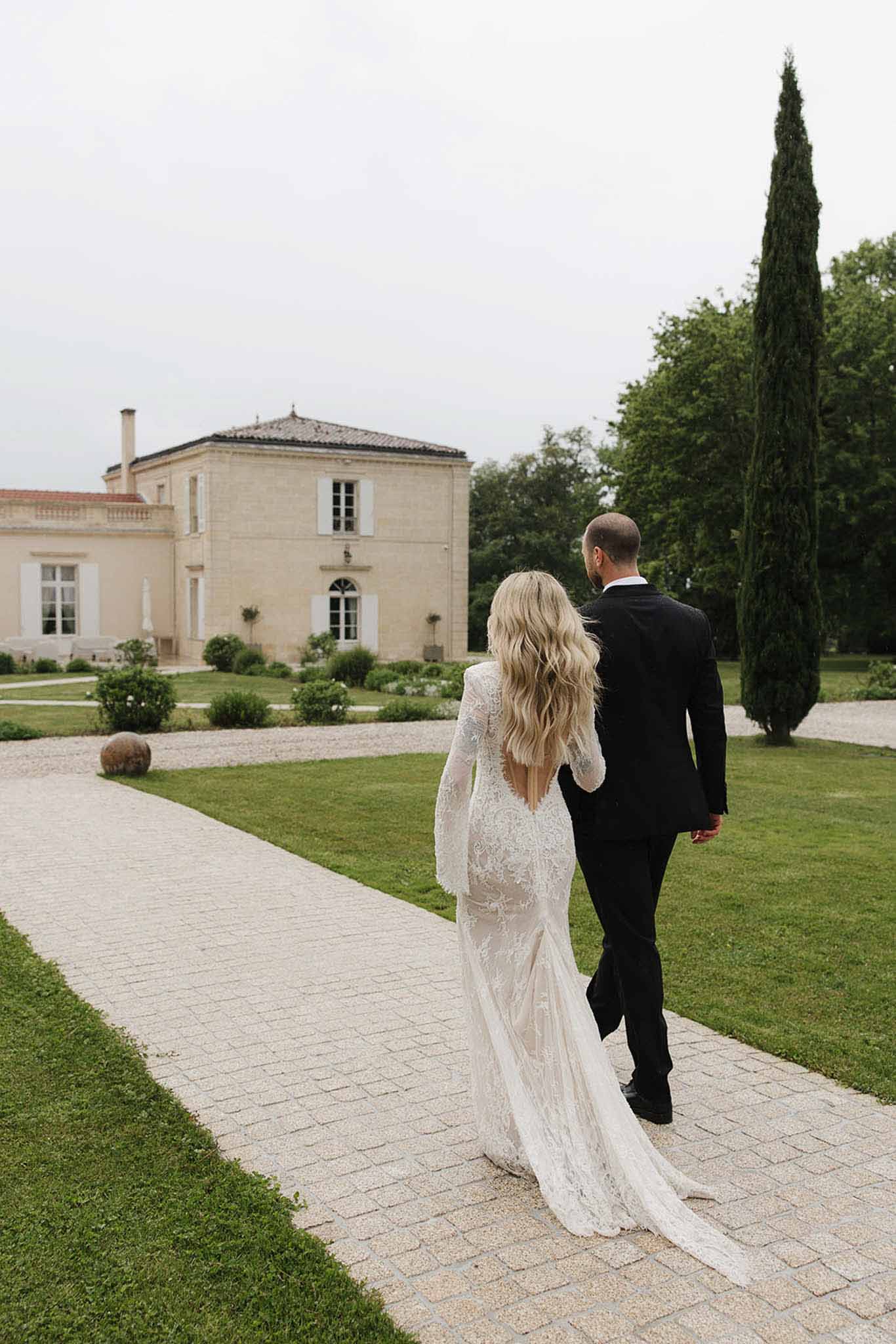 Bride and groom walking away toward limestone chateau on gravel path with manicured hedges