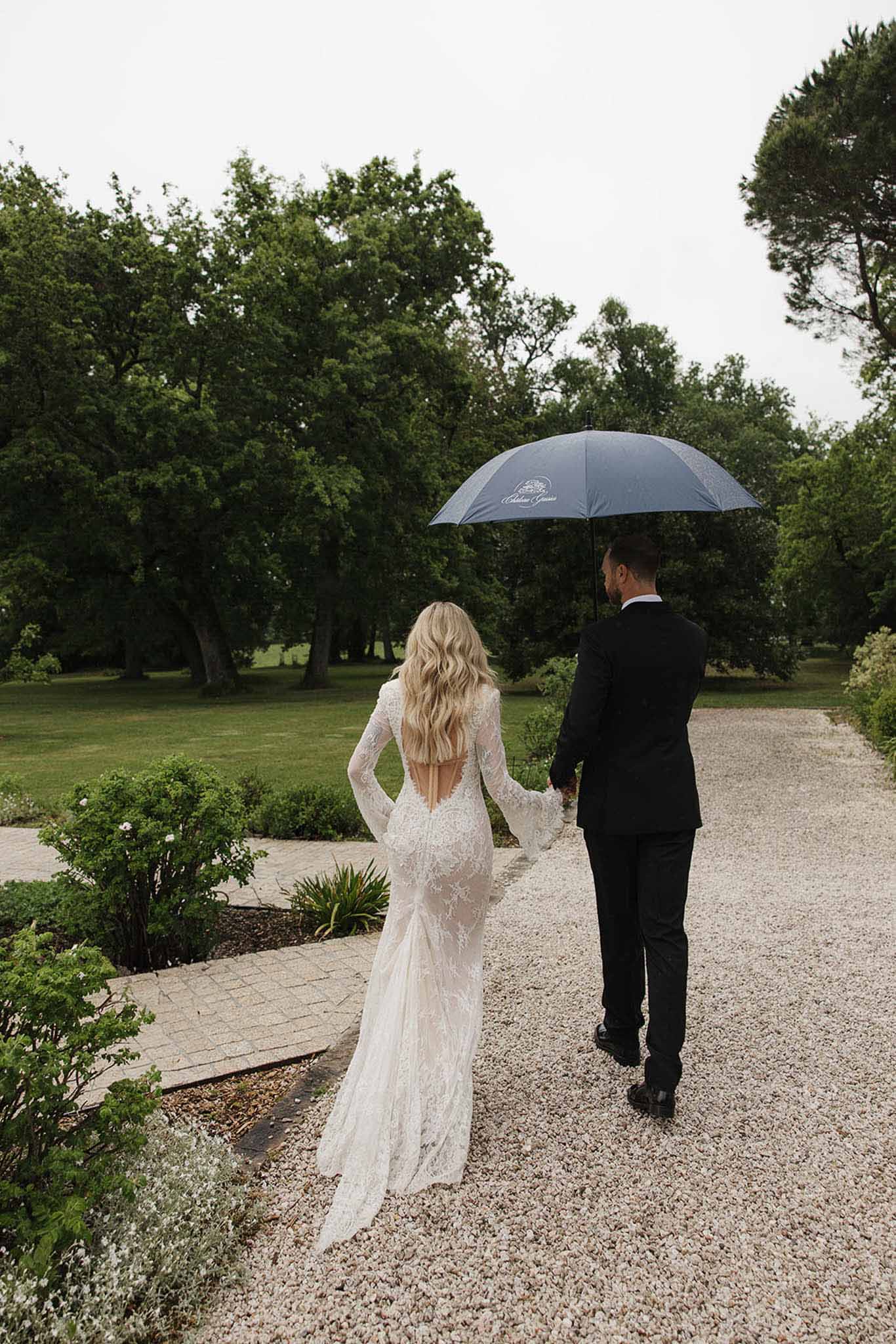 Couple walking from behind on rainy day under branded umbrella, bride in open-back ivory lace gown on chateau grounds
