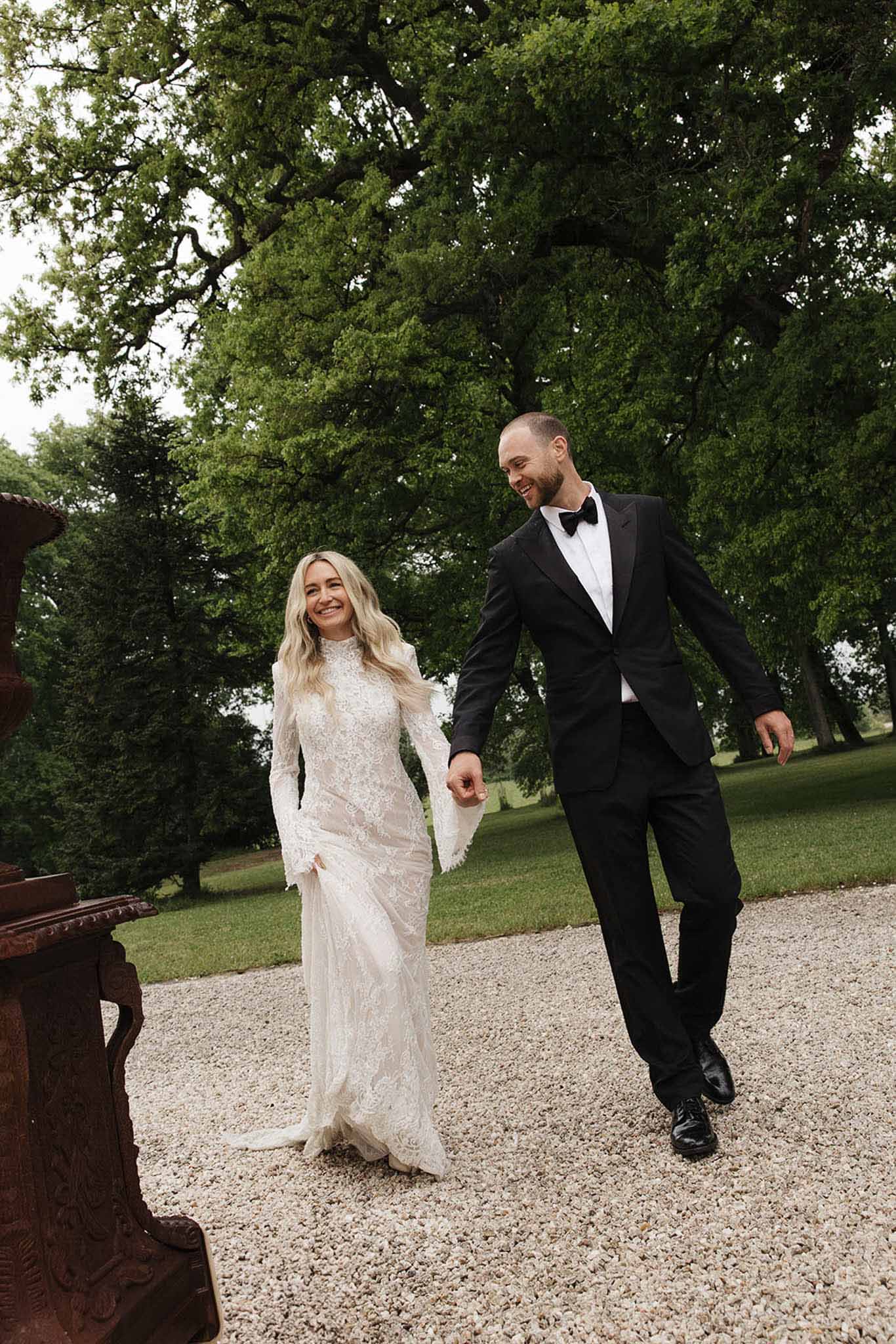 Couple laughing and walking on gravel path with bride in long-sleeve lace gown at chateau estate