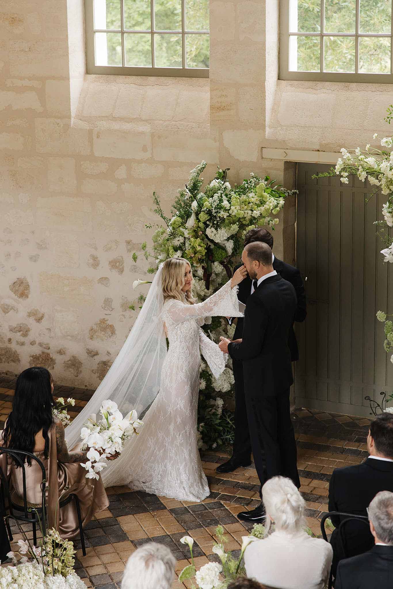 Bride in lace gown wiping tear from groom's face at altar framed by white calla lilies in a stone chapel