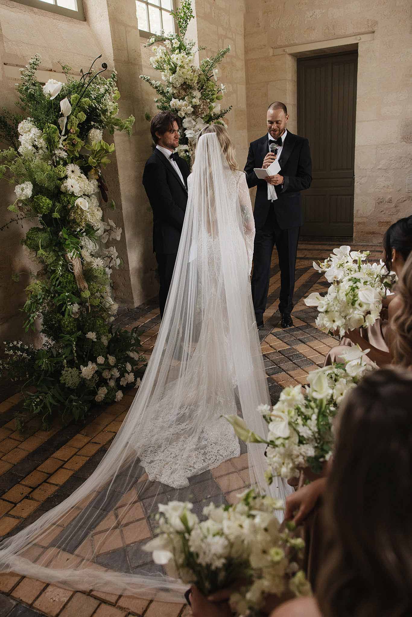 Ceremony from behind with calla lily and orchid altar installation, lace cathedral veil, and bridesmaids with bouquets