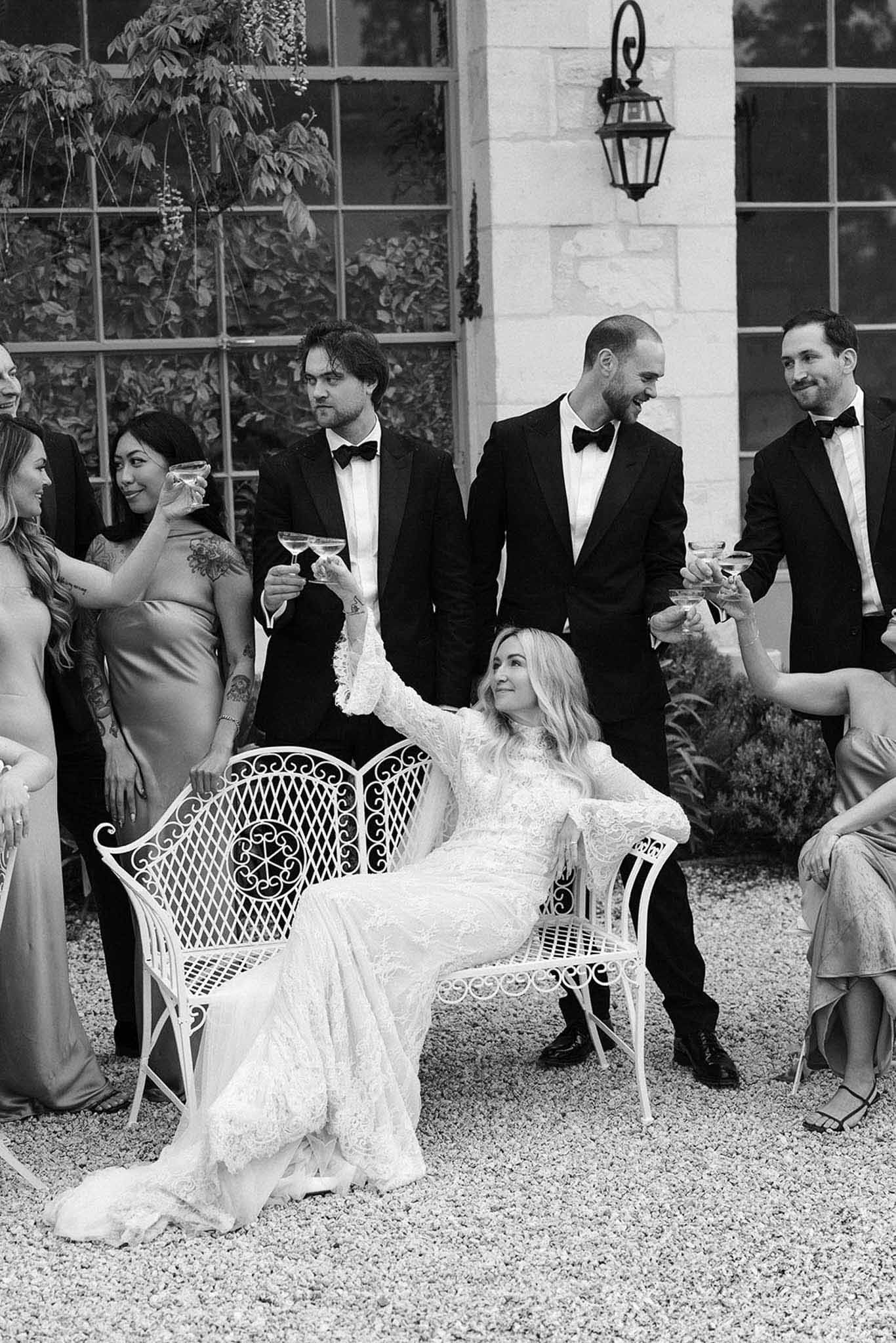 Bride on wrought-iron bench toasting with bridesmaids and groomsmen in chateau courtyard in B&W