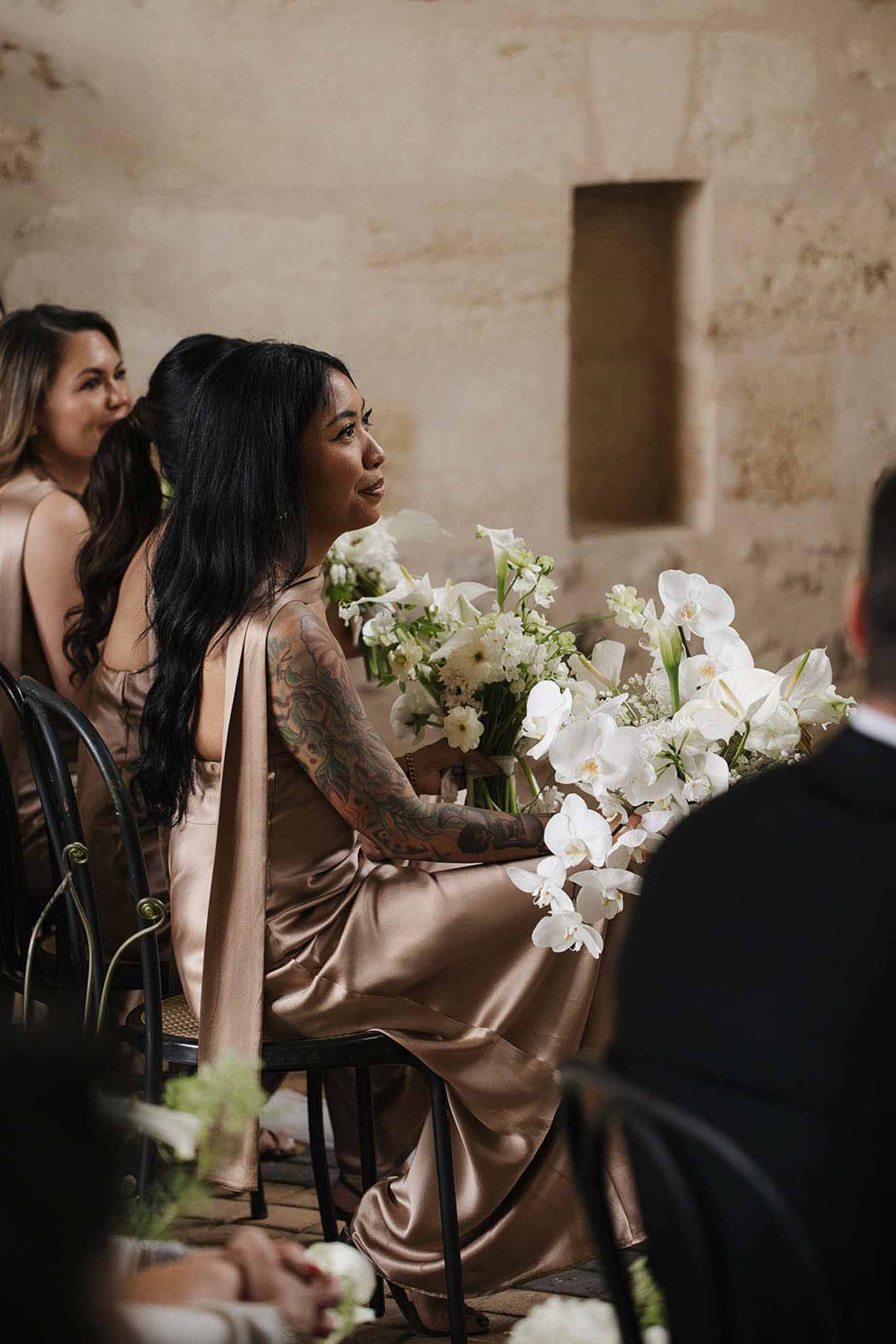 Bridesmaid in champagne satin dress holding white orchid and calla lily bouquet during ceremony