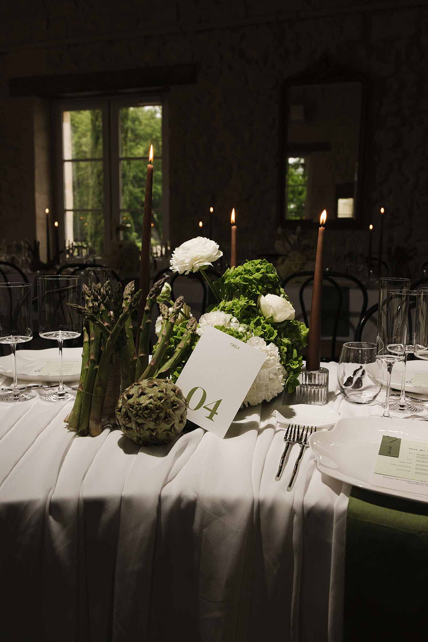 Reception table centerpiece with artichoke, asparagus, white ranunculus, and brown taper candles beside a green table numb...