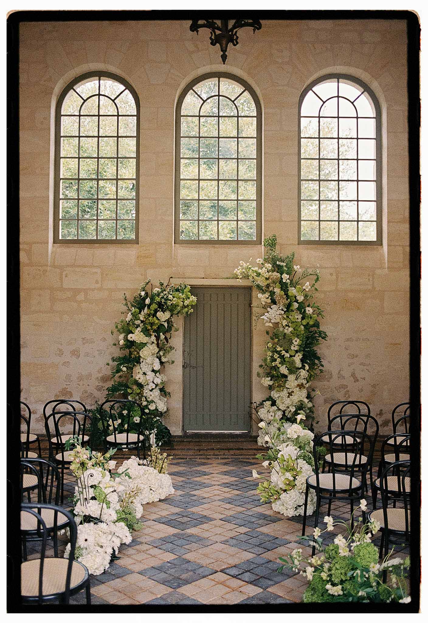 Chapel ceremony setup with white hydrangea arch around sage door, bentwood chairs, and diamond-tile floor