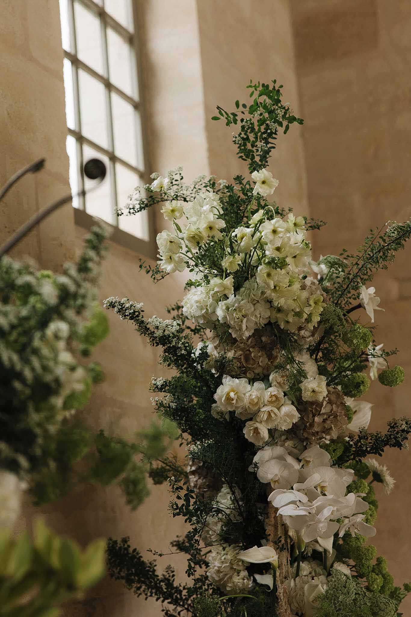 Lush white ceremony installation of hydrangeas, orchids, calla lilies, and trailing greenery in stone chapel