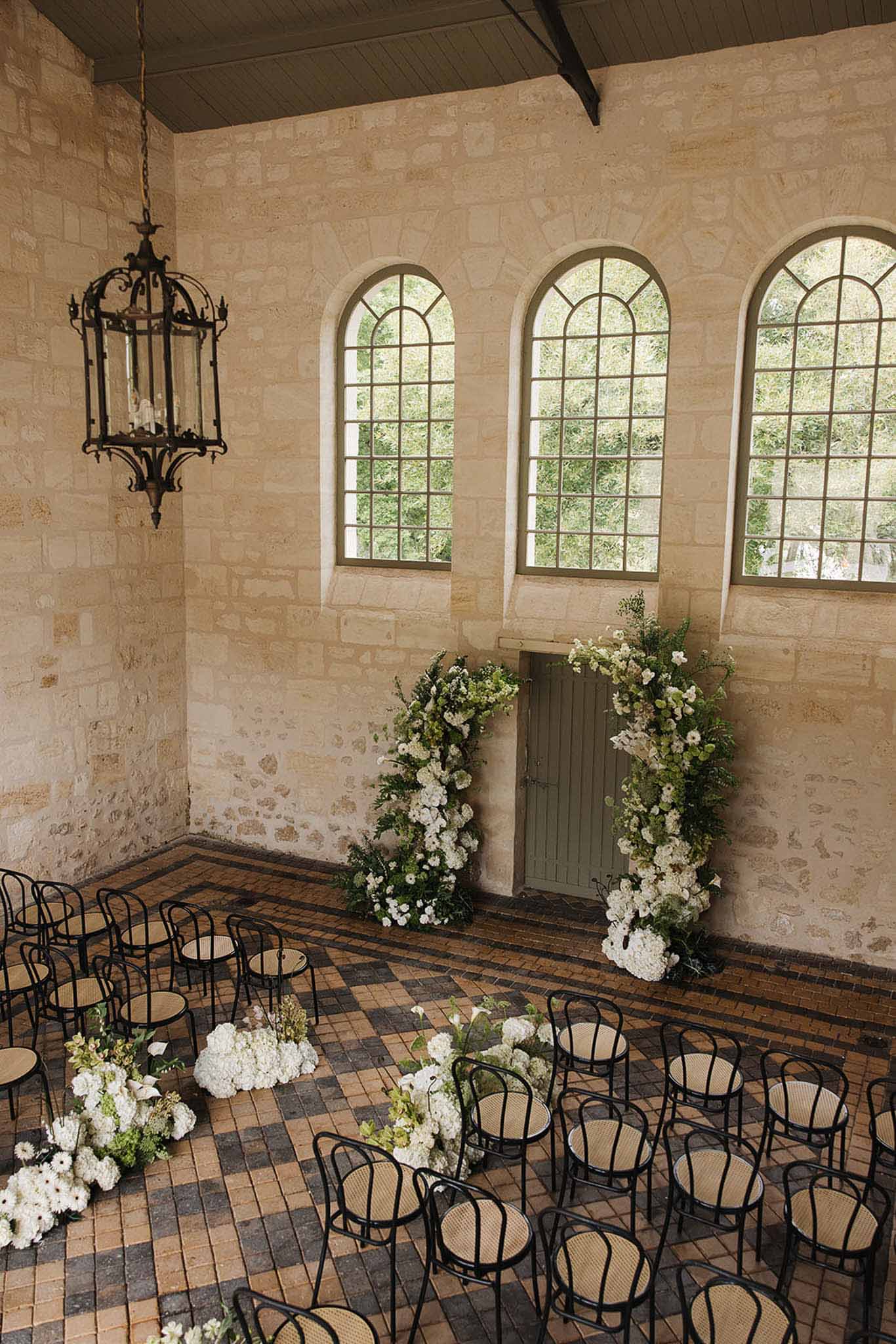 Indoor ceremony with white hydrangea and rose arch framing sage green door in limestone hall from above