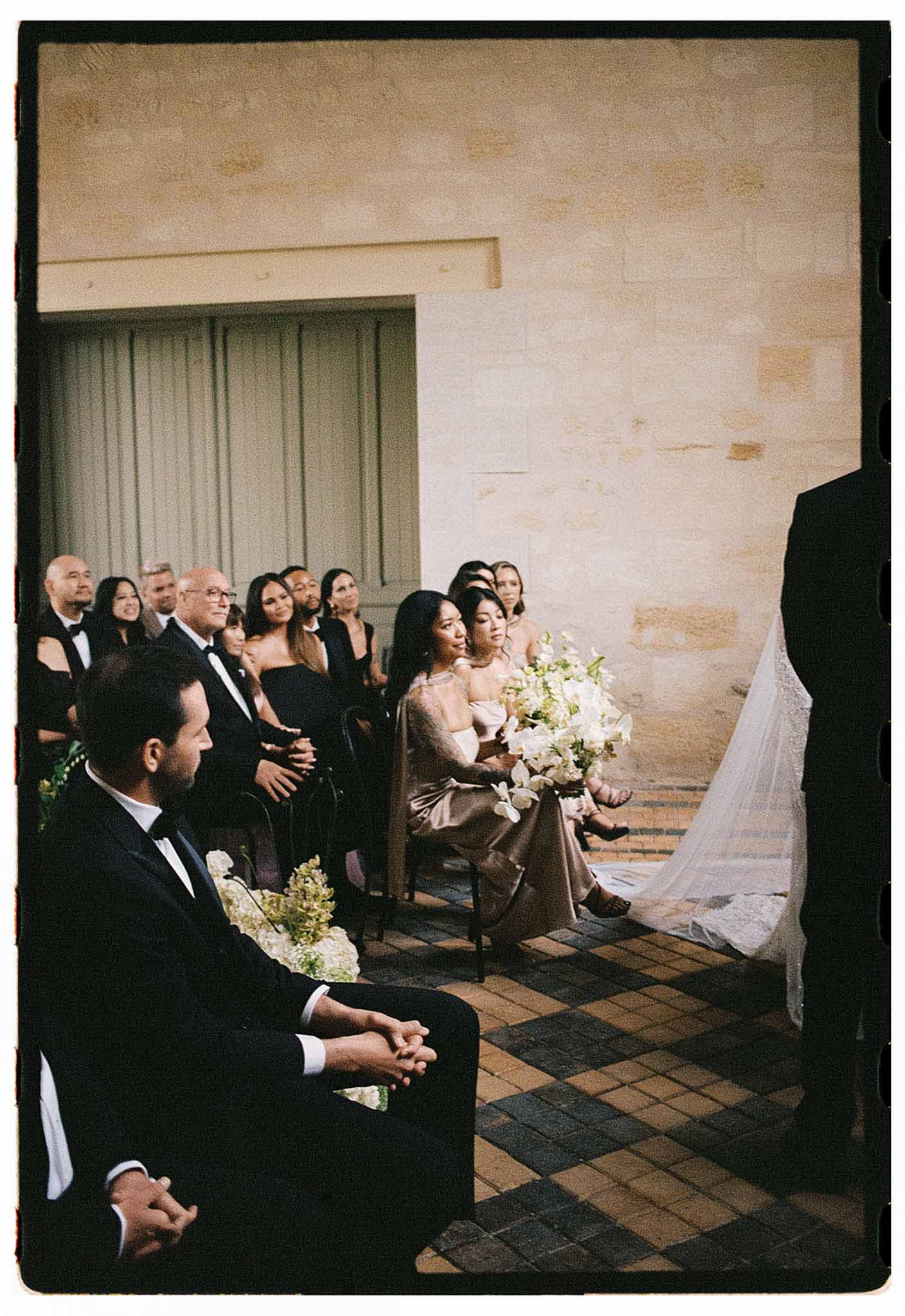Film-style wide shot of outdoor wedding ceremony with seated guests, bridesmaids in champagne dresses holding white bouquets