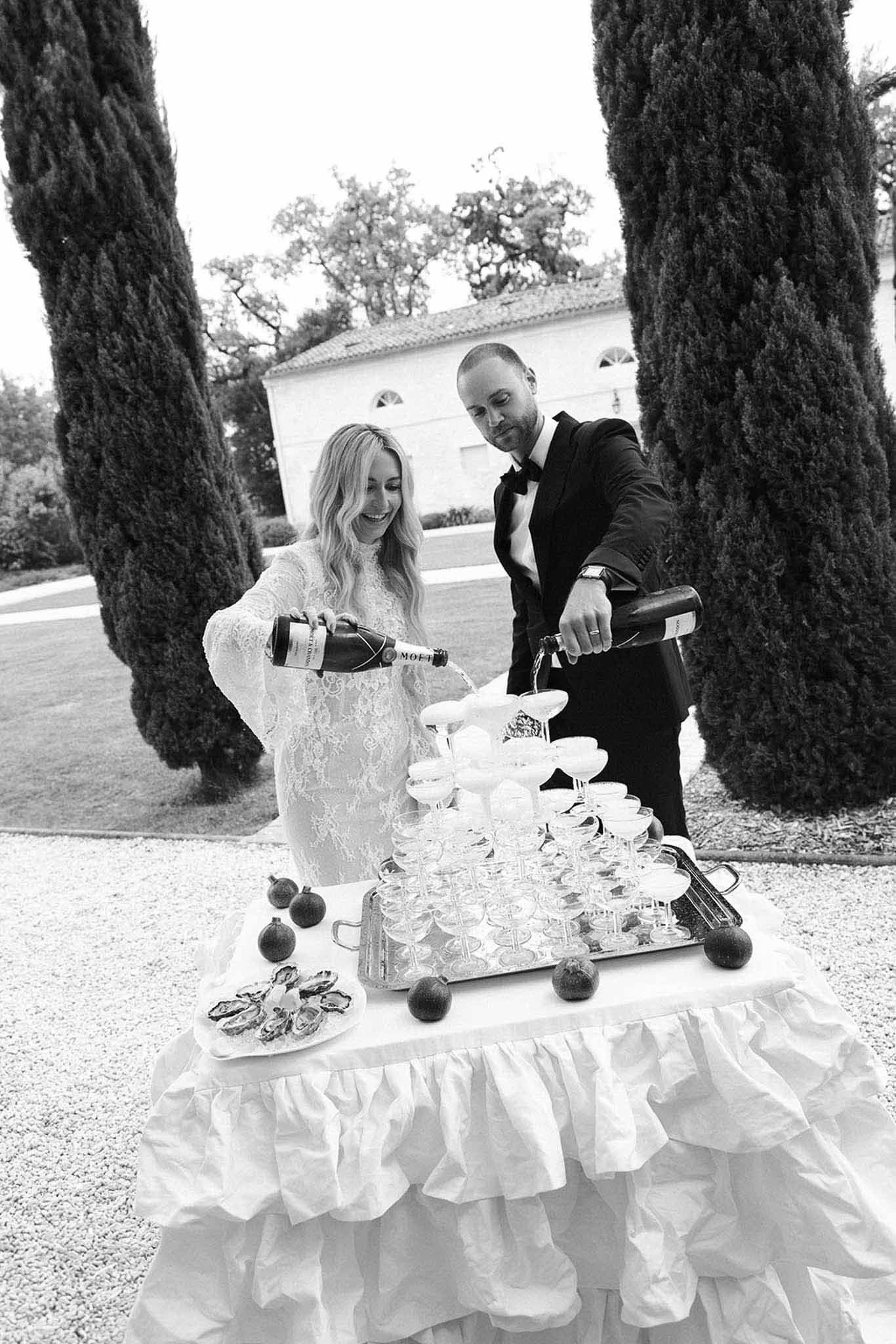 Couple pours Moet into coupe tower beside oysters and figs on ruffled linen before cypress trees in B&W