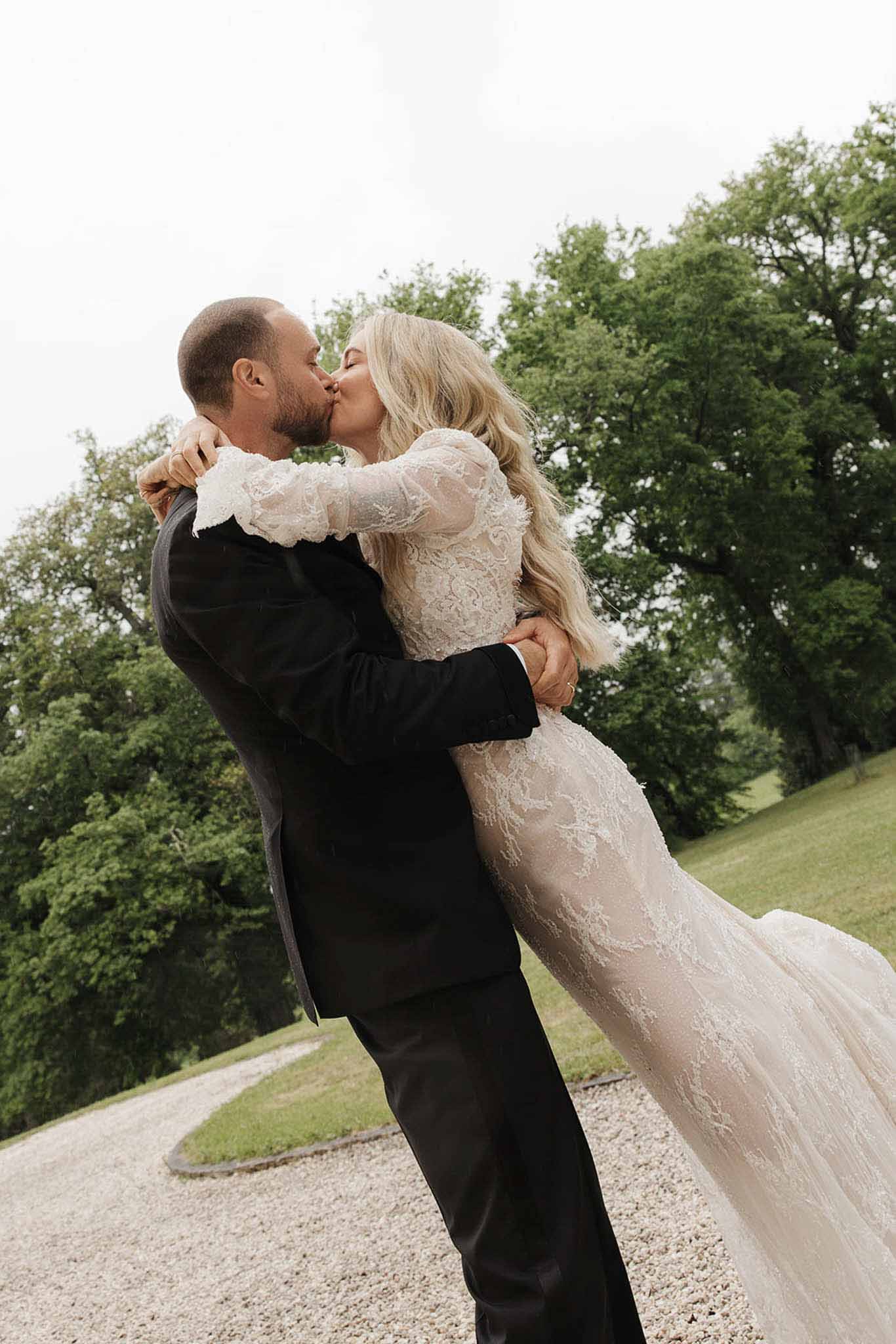 Groom lifting bride off ground for kiss in long-sleeve lace gown on estate driveway